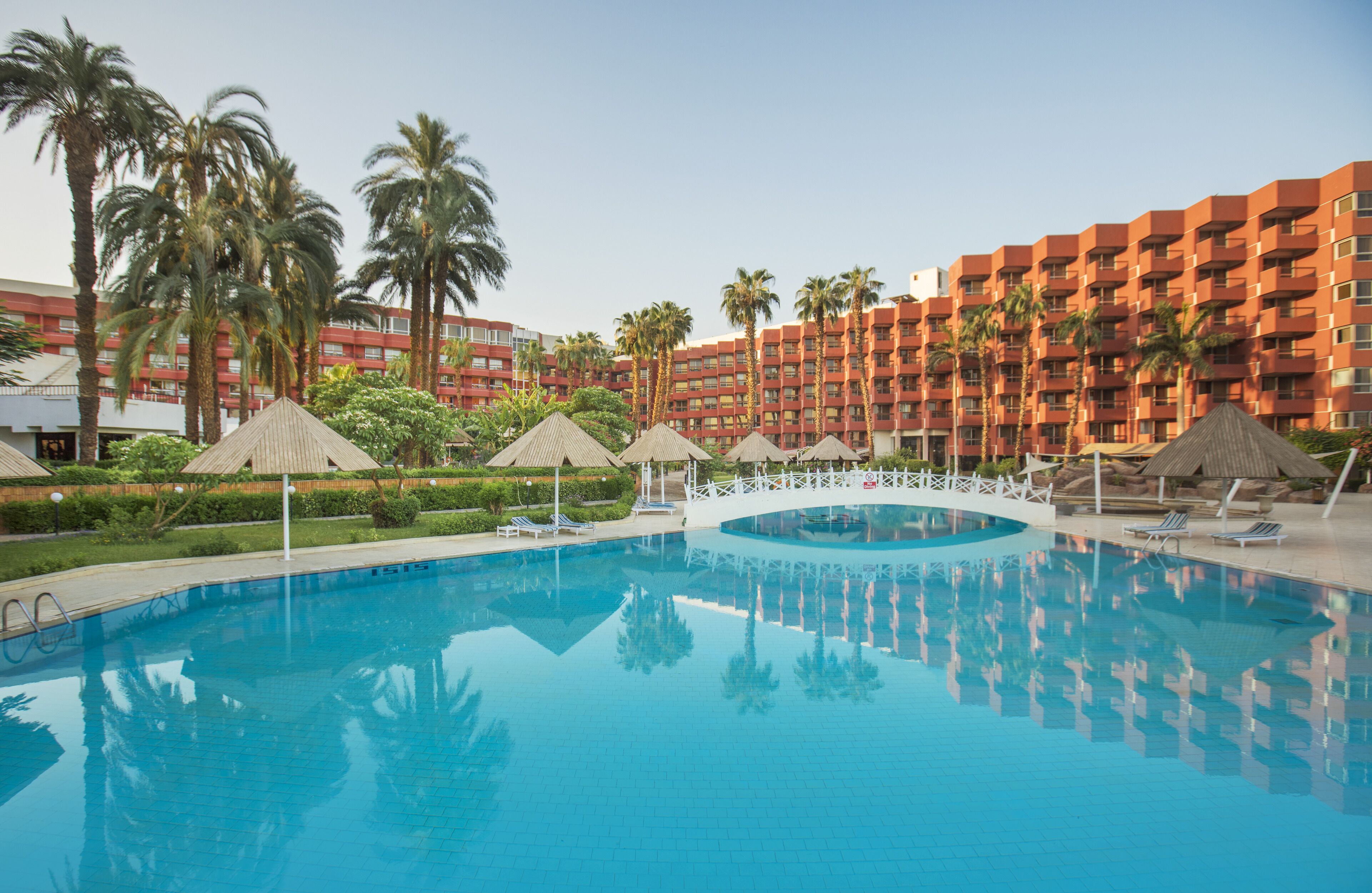 A swimming pool with lounge chairs and umbrellas, surrounded by palm trees and a red building with balconies.