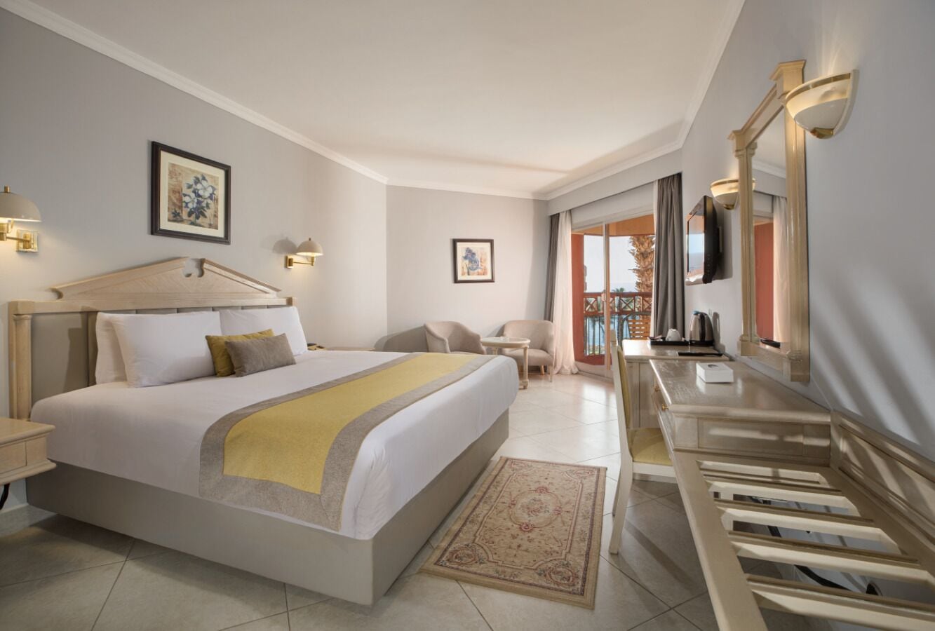A modern hotel room with a large bed, a sitting area with a chair, a desk, and a TV mounted on the wall.