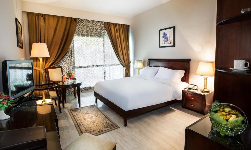 A hotel room with a bed, bedside tables, a desk with a lamp, a television, and a window with curtains.