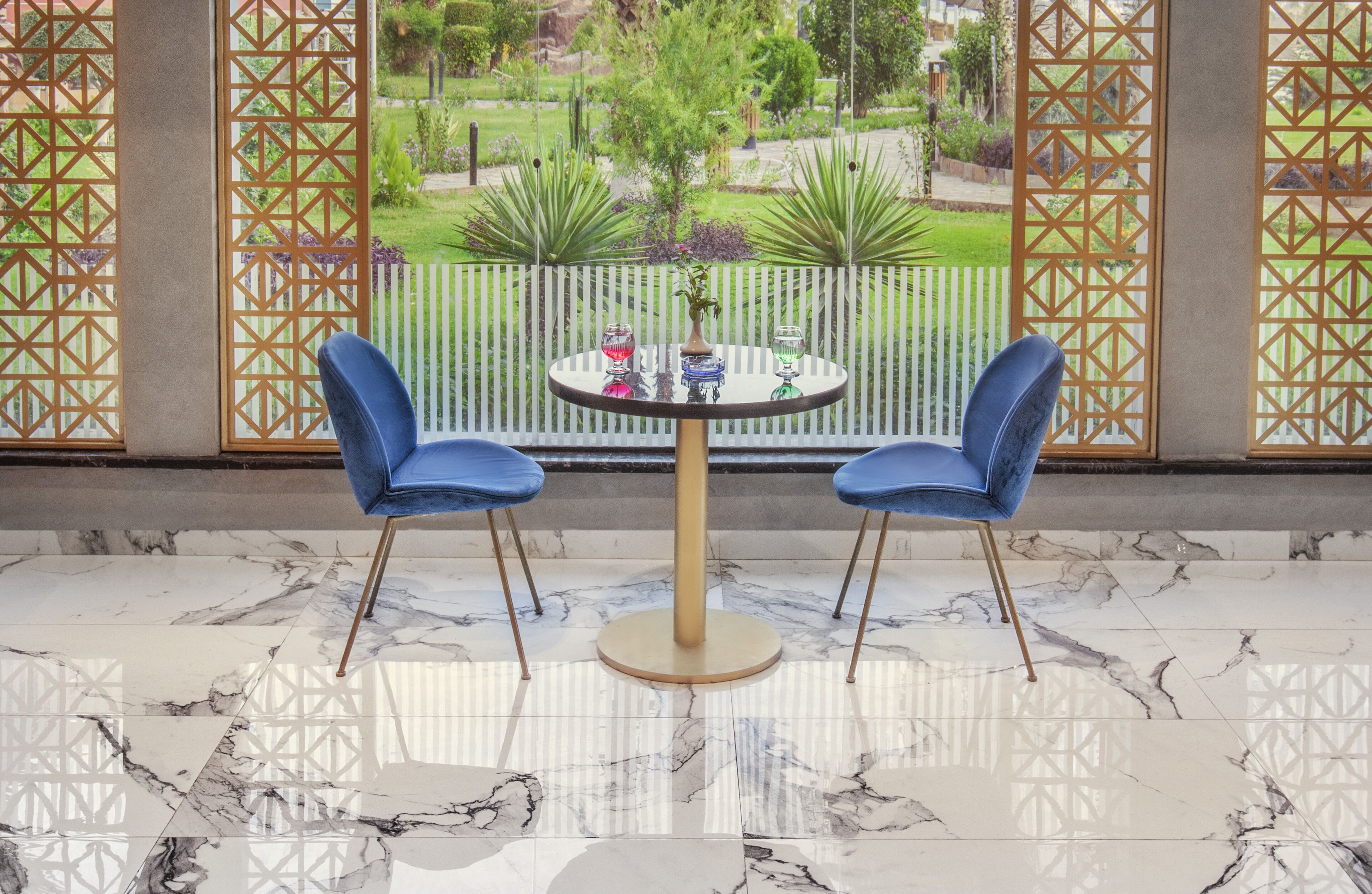 Una habitación con piso de mármol, dos sillas azules, una mesa redonda con un florero y vista a un jardín a través de una ventana con celosías.