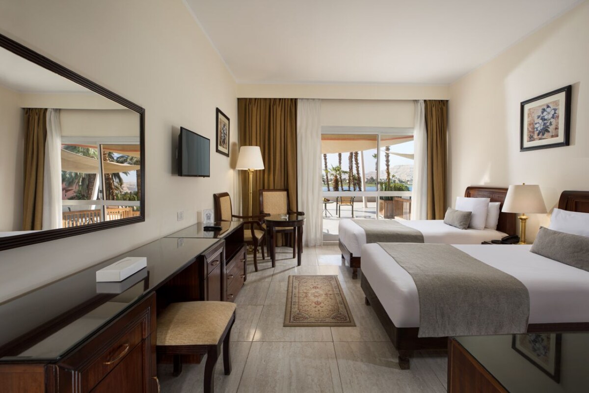 A hotel room with a desk, two beds, a chair, a TV, and a balcony with a view.