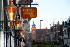 A hotel sign with the name "HOTEL OORSPROONG PARK" and a view of a street with traditional buildings and a clock tower in the background.