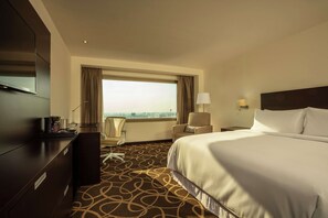 A hotel room with a large bed, a desk with a chair, a TV, a window with curtains, and a view of the city.