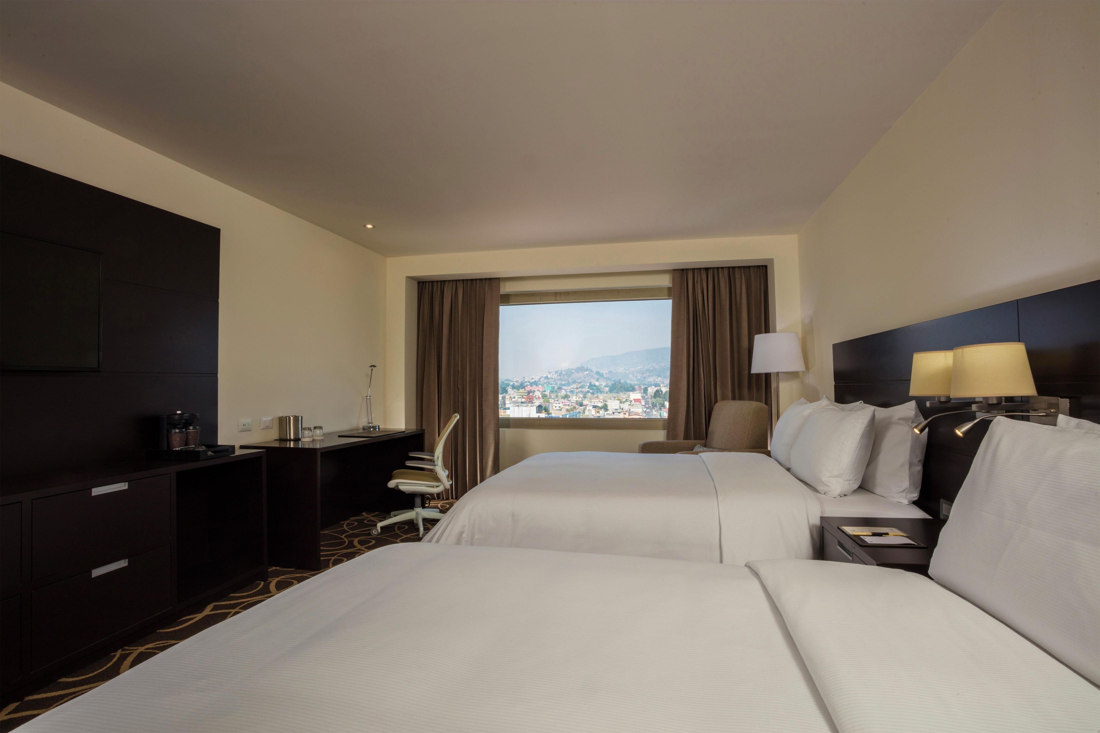 A hotel room with two beds, a desk, a chair, and a view of a cityscape.