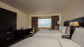 A hotel room with two beds, a desk, a chair, and a view of a cityscape.