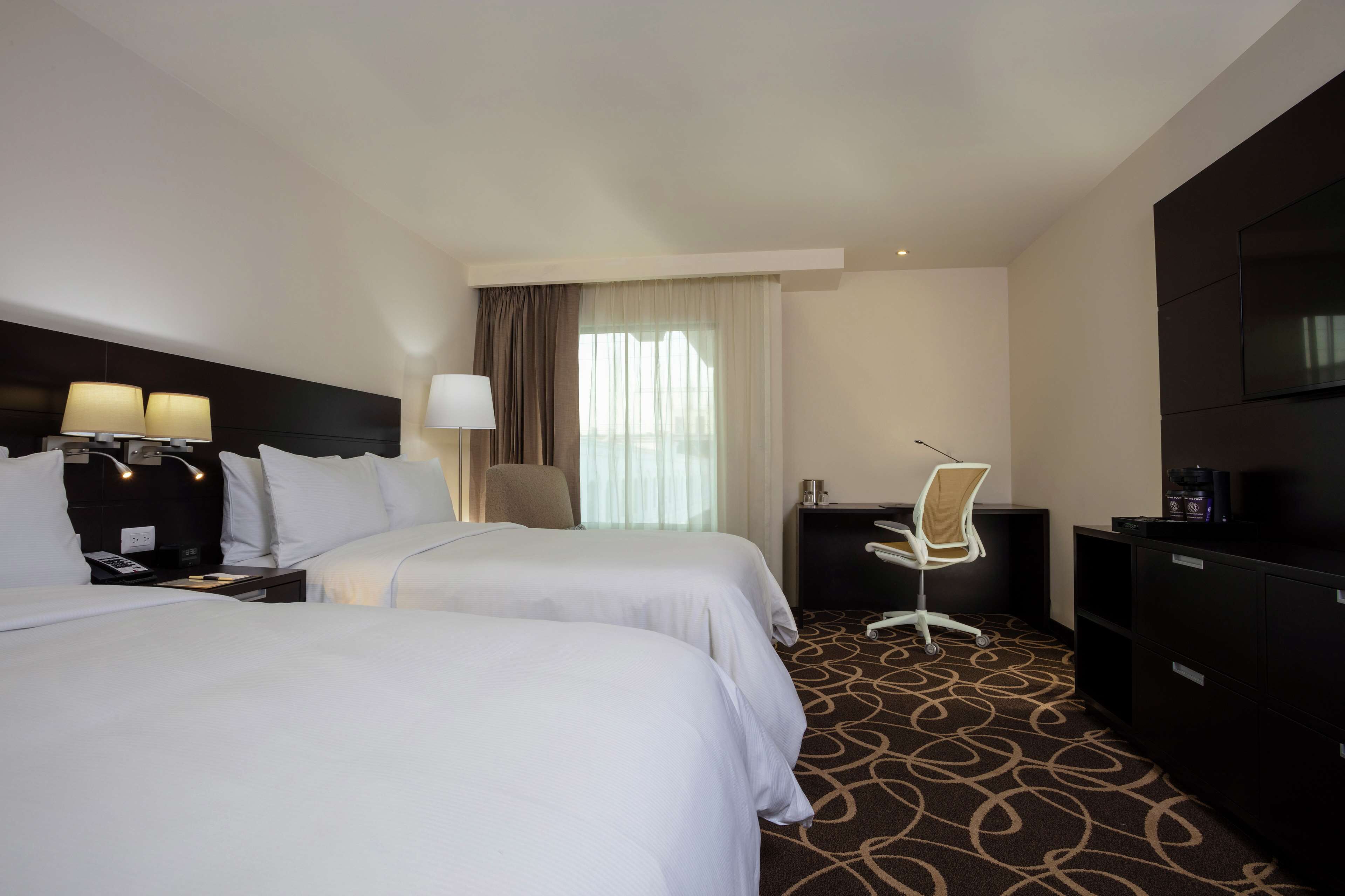 A hotel room with two beds, a desk, a chair, a television, and a window with curtains.