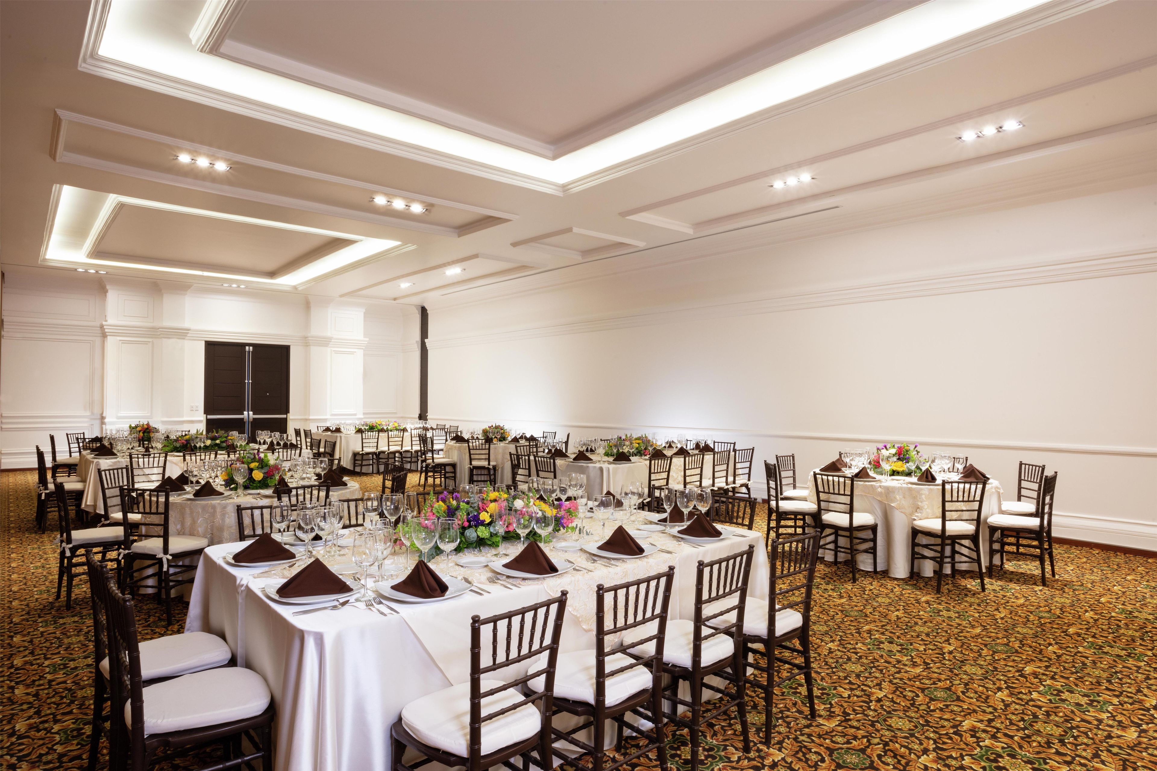 Un salón de banquetes con varias mesas redondas dispuestas para comer, cada una con mantel blanco y servilletas marrones.