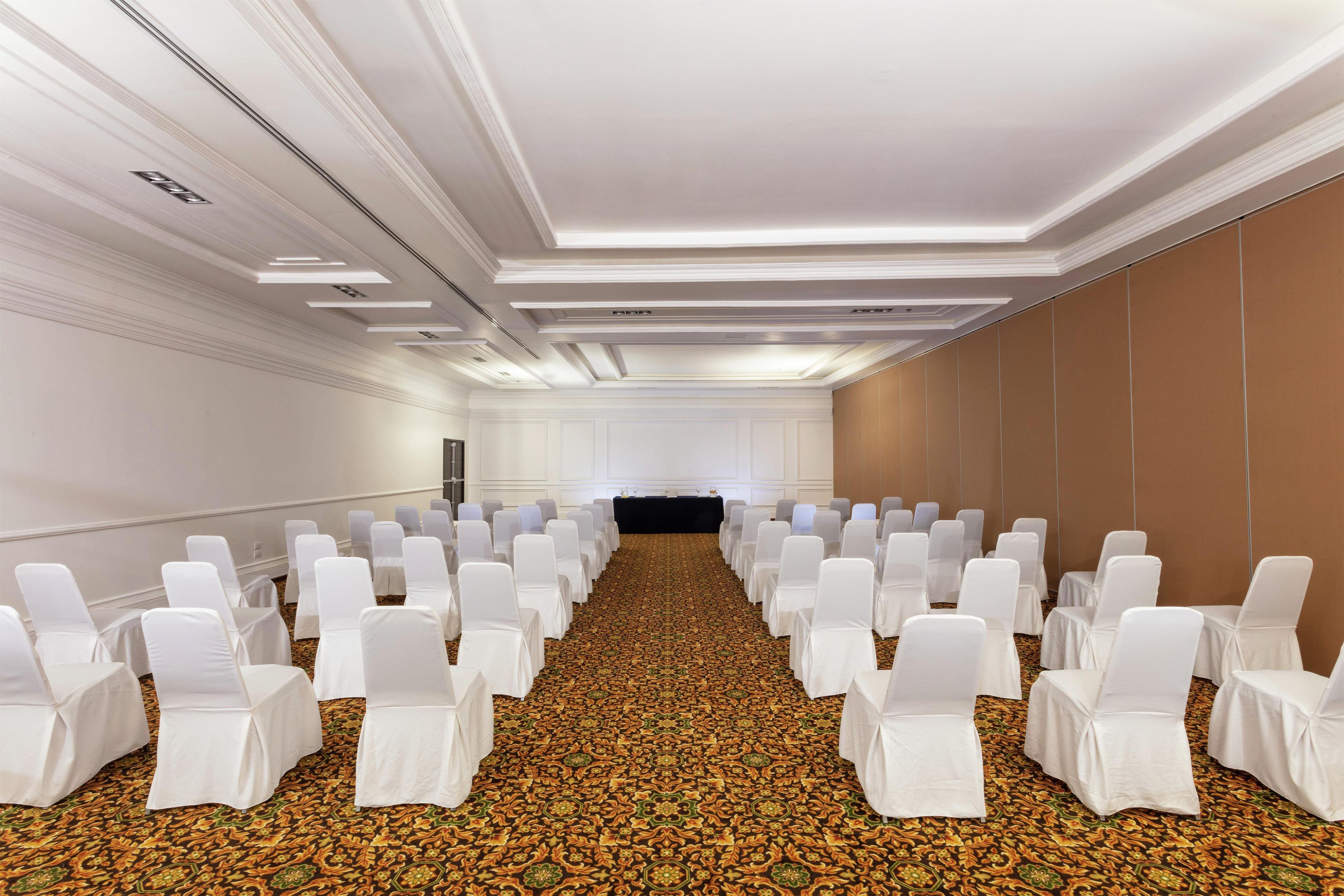 A conference room with rows of white chairs and tables, a patterned carpet, and a plain wall with a door at the far end.