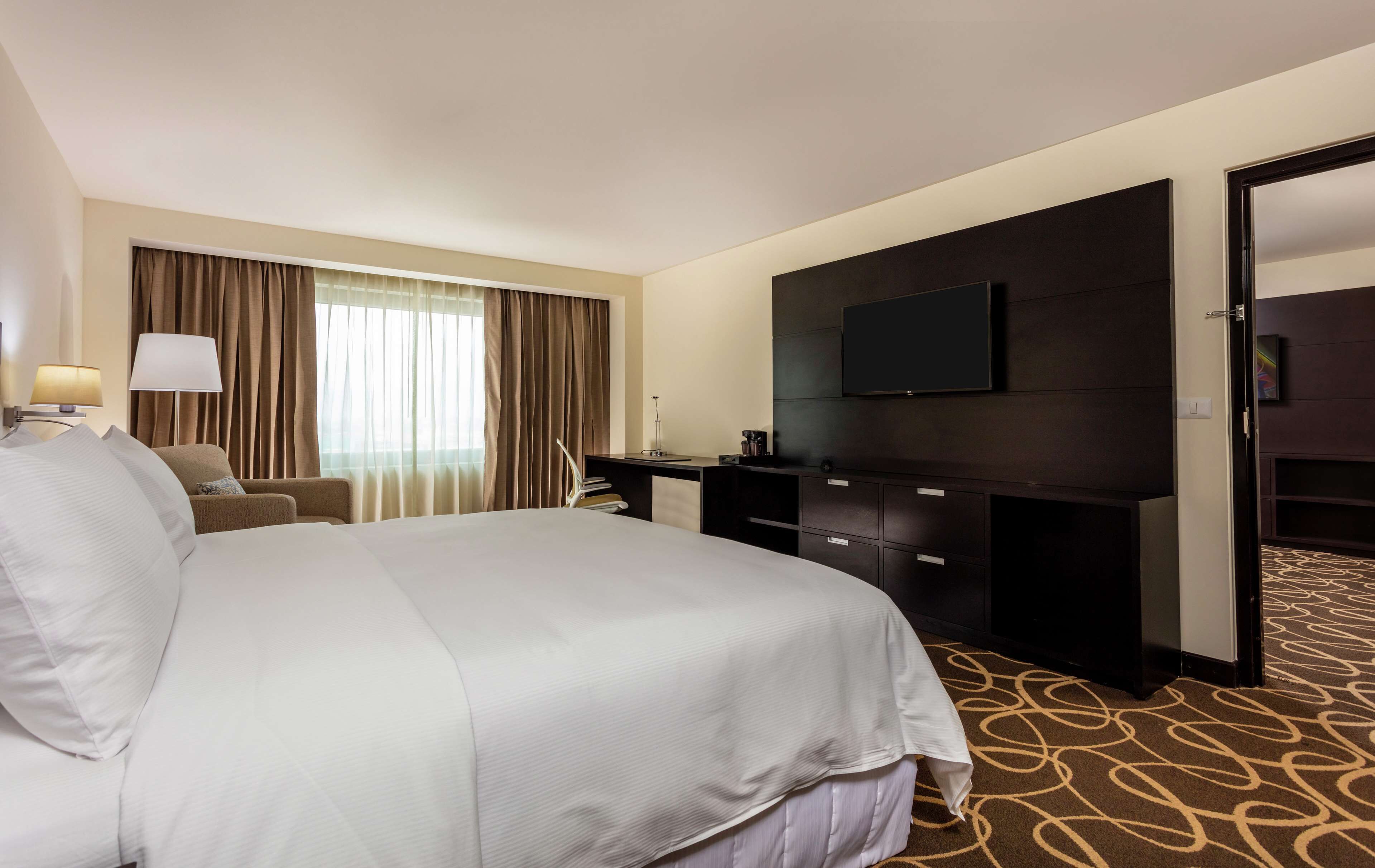 A hotel room with a large bed, a desk with a flat-screen TV, a chair, and a desk lamp.