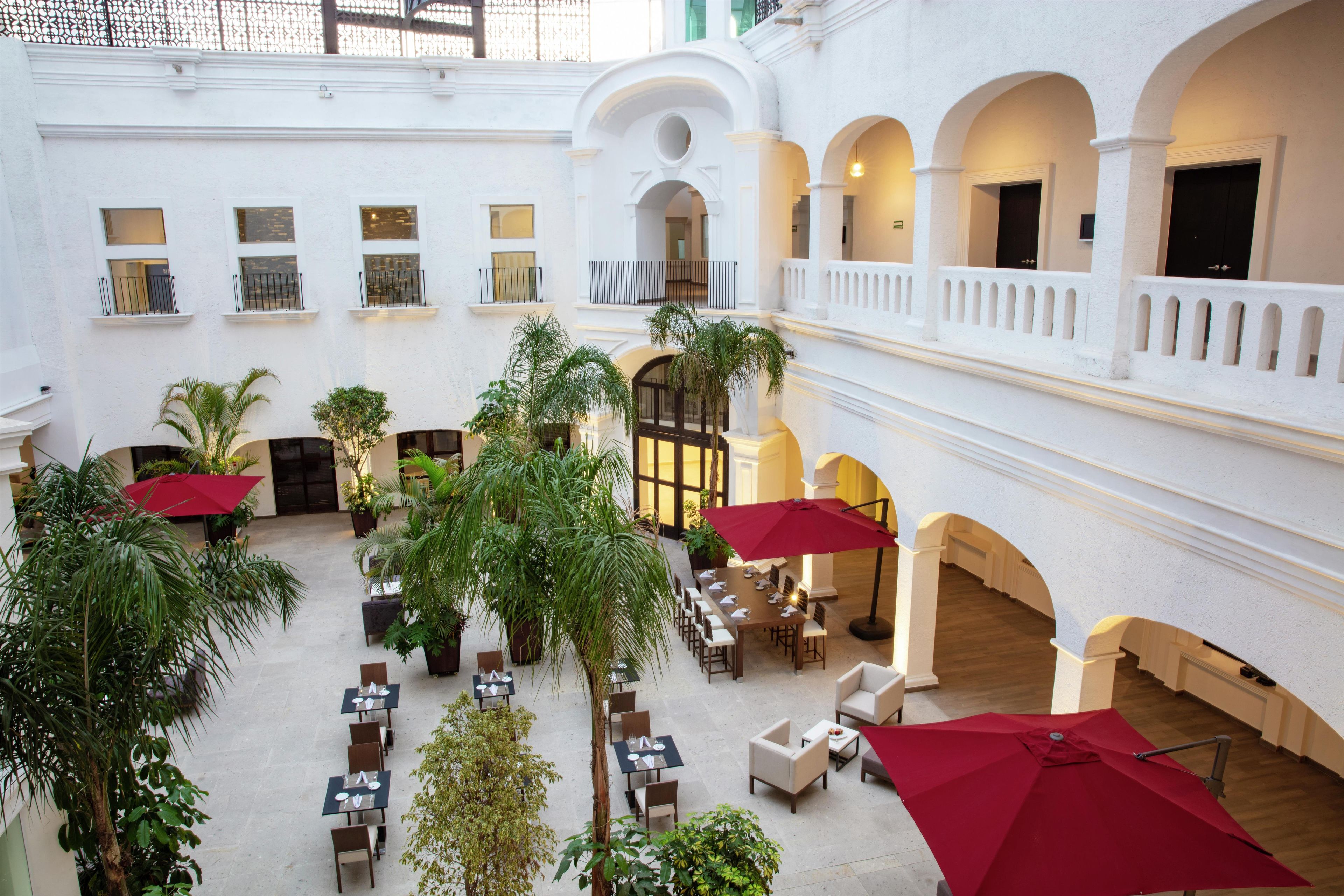 A spacious indoor courtyard with multiple dining tables and chairs, surrounded by lush greenery and red umbrellas.
