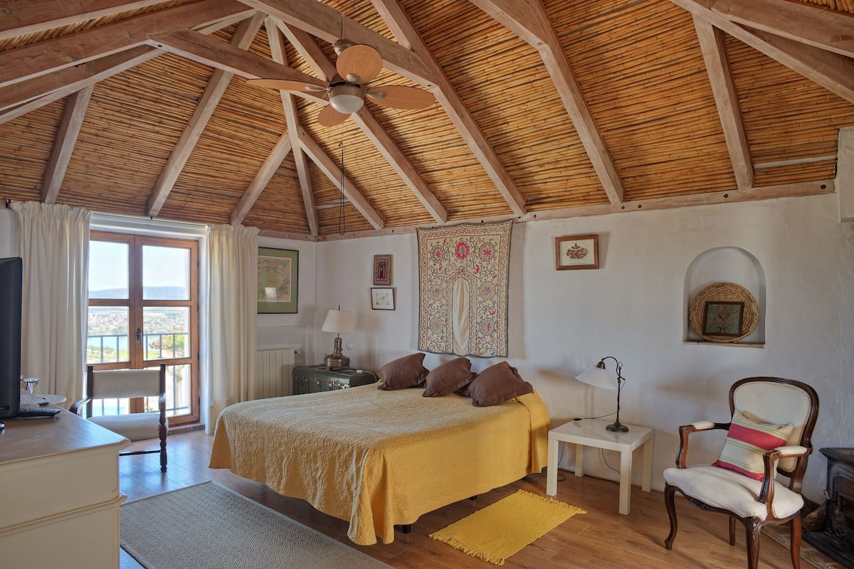 Ein Schlafzimmer mit Holzbalkendecke, einem Bett mit gelber Bettdecke, einem Sessel, einem Schreibtisch und einem Fenster mit Vorhängen.