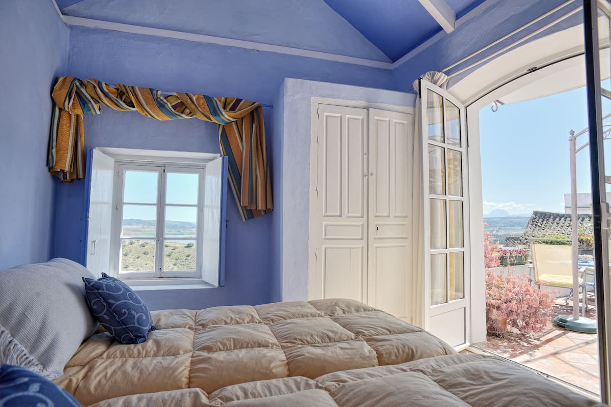 Ein Schlafzimmer mit Bett, Fenster, Tür und Blick ins Freie.