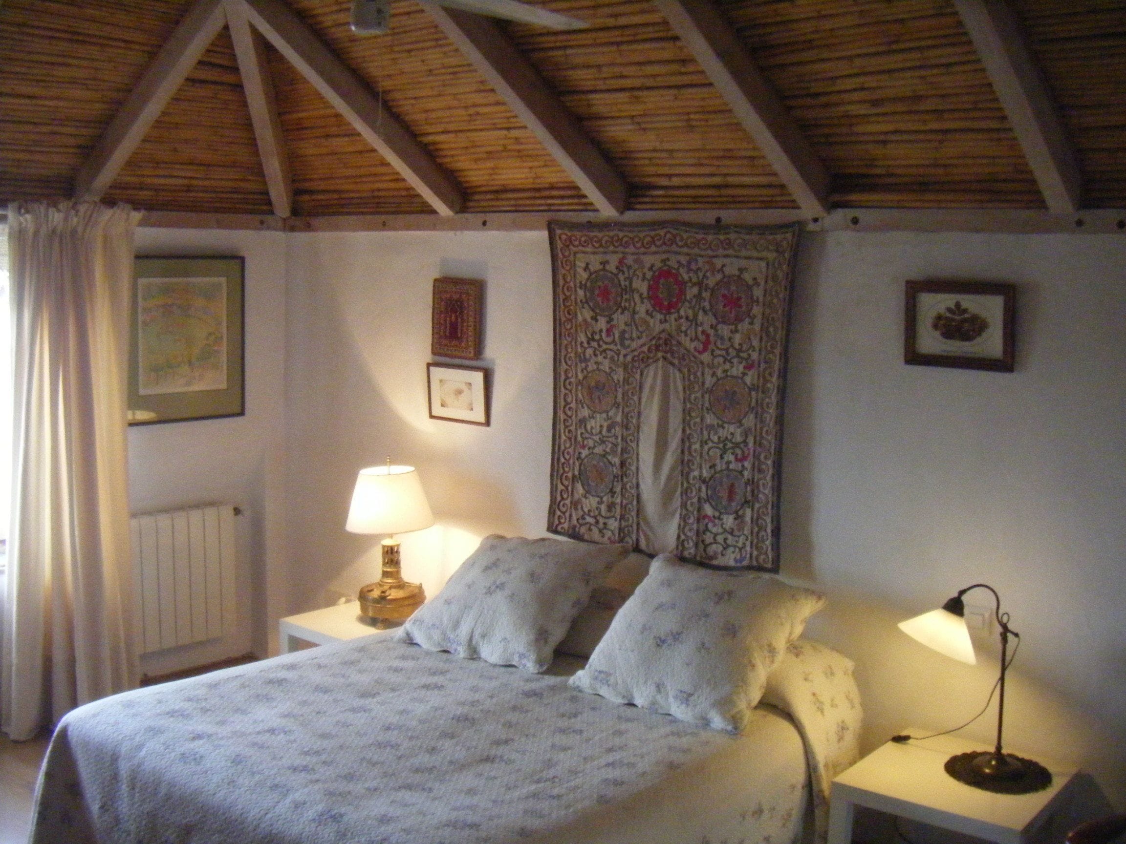 A bedroom with a wooden ceiling, a bed with floral bedding, two bedside tables with lamps, and a tapestry on the wall.