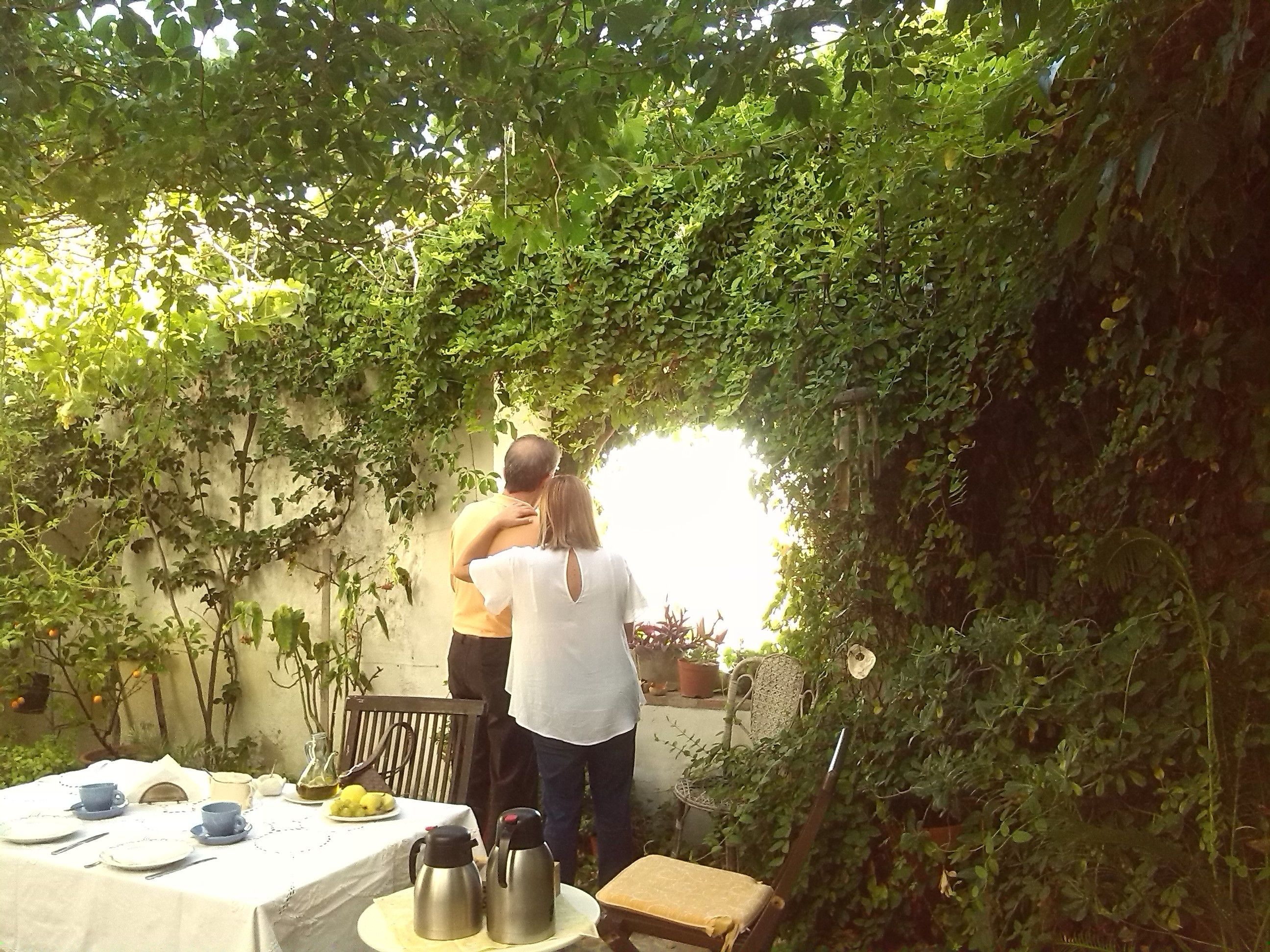 A couple embracing under a leafy arbor in a garden setting with a table set for two, including cups, plates, and a kettle.