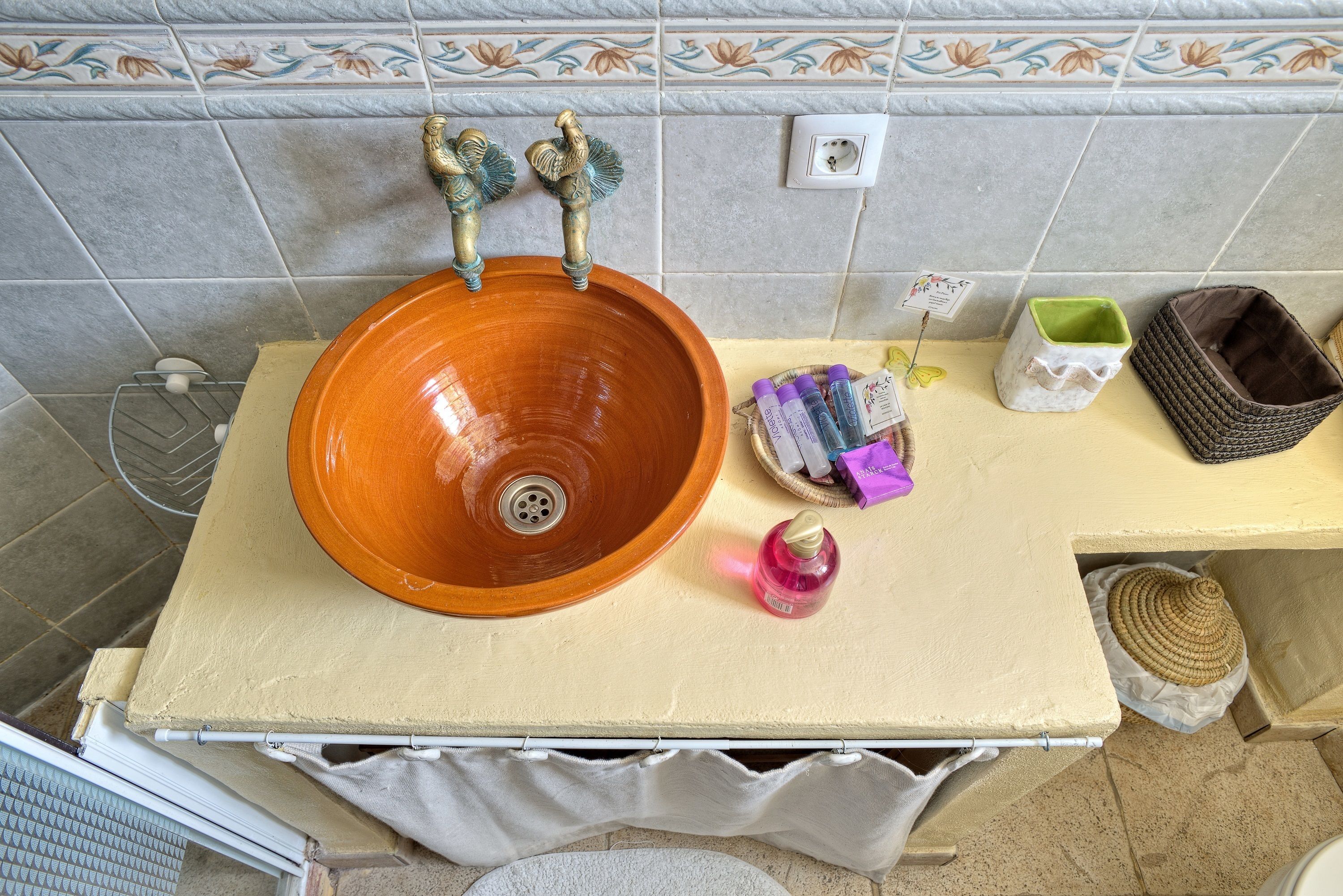 A bathroom with a large, round, orange sink, two faucets, a small white container, a woven basket, and a towel rack.