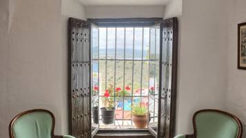 A corner room with two green upholstered chairs, a small round table, and a large window with a view of a garden and mountains.