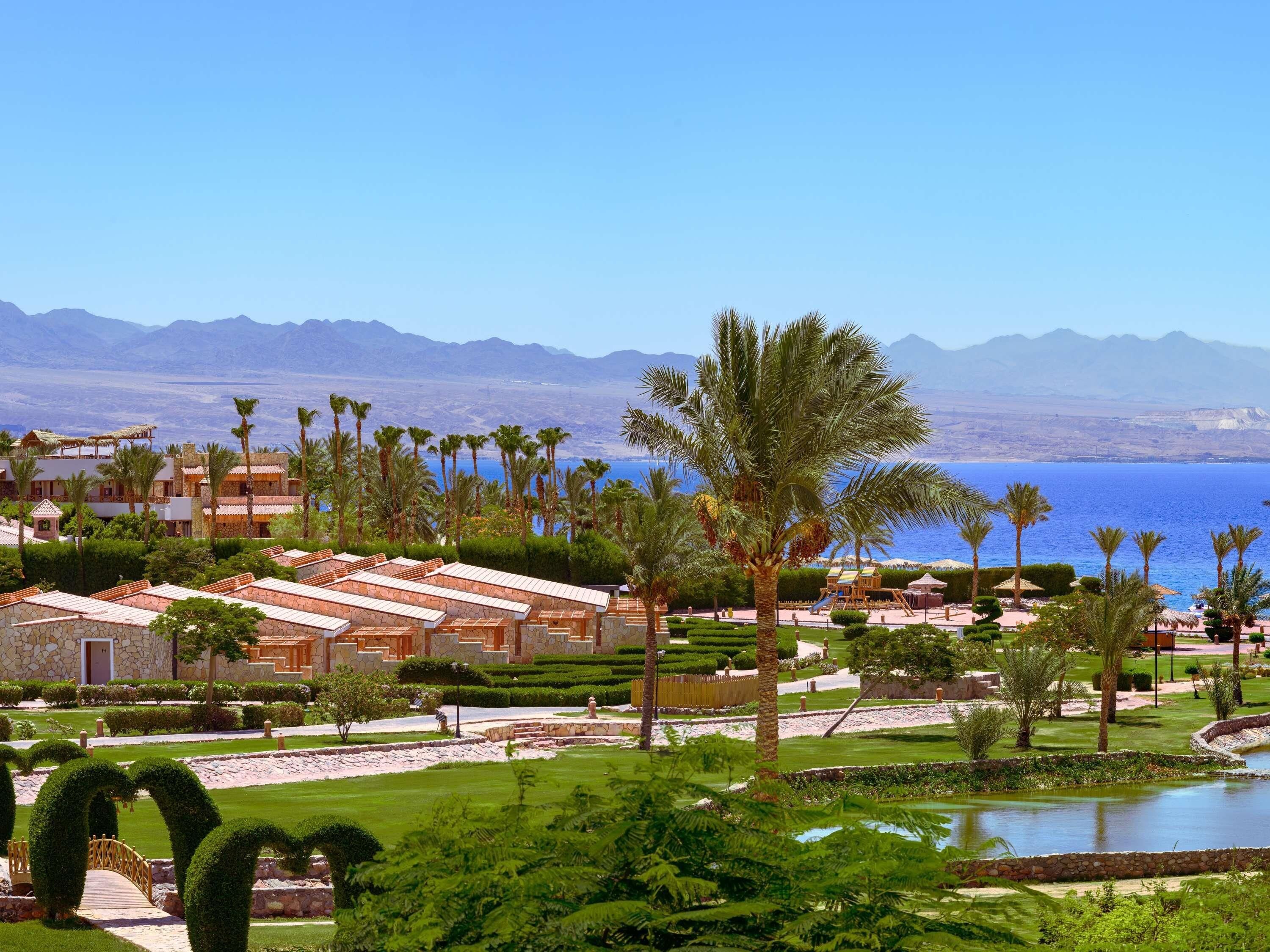 Un resort con varias construcciones, palmeras y vistas al mar y las montañas.