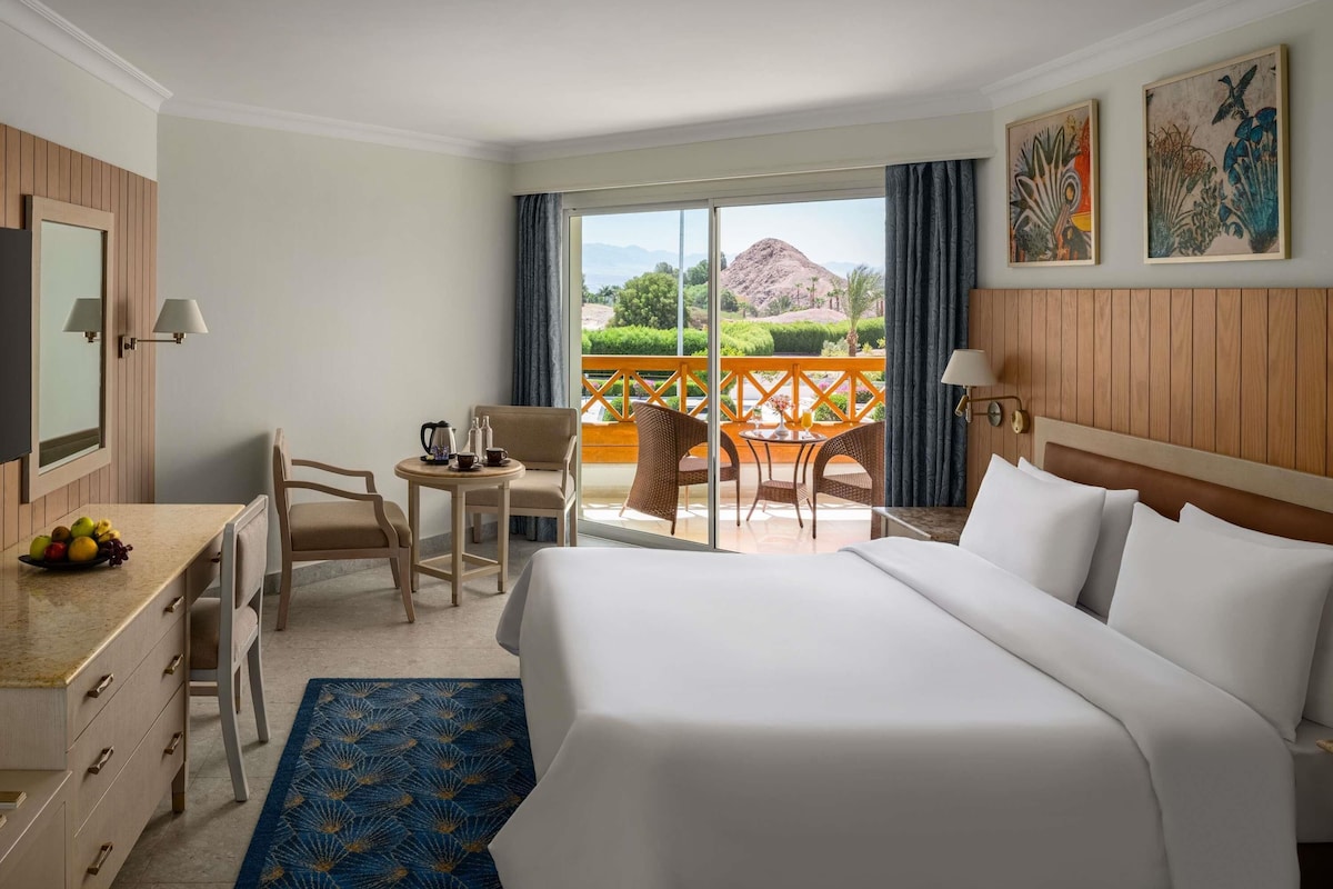 A hotel room with a bed, a desk with a fruit bowl, a chair, a small table with a coffee pot, and a balcony with a view of a mountain and greenery.