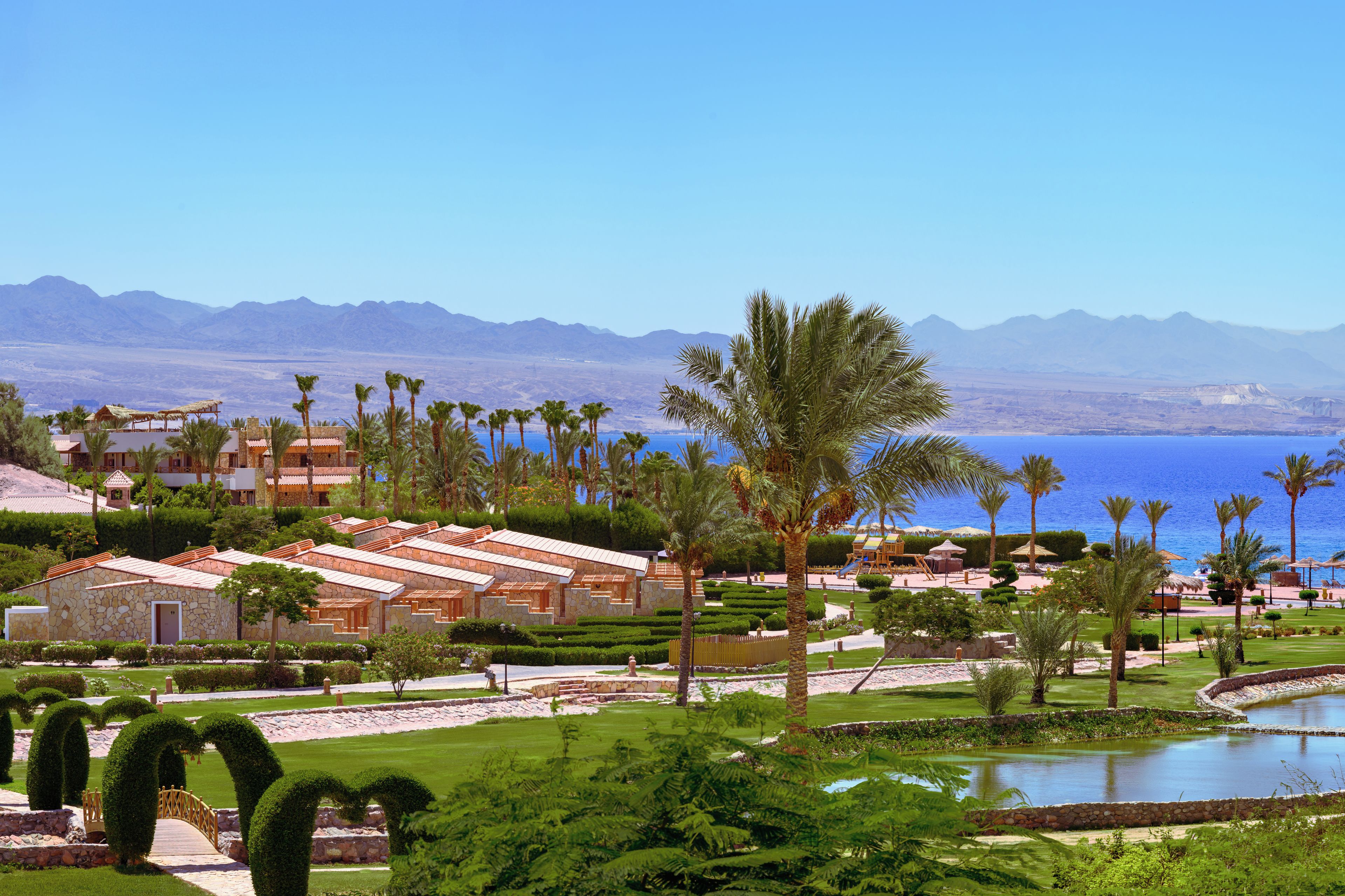 Un resort con varias construcciones, palmeras, una piscina y vistas a las montañas.