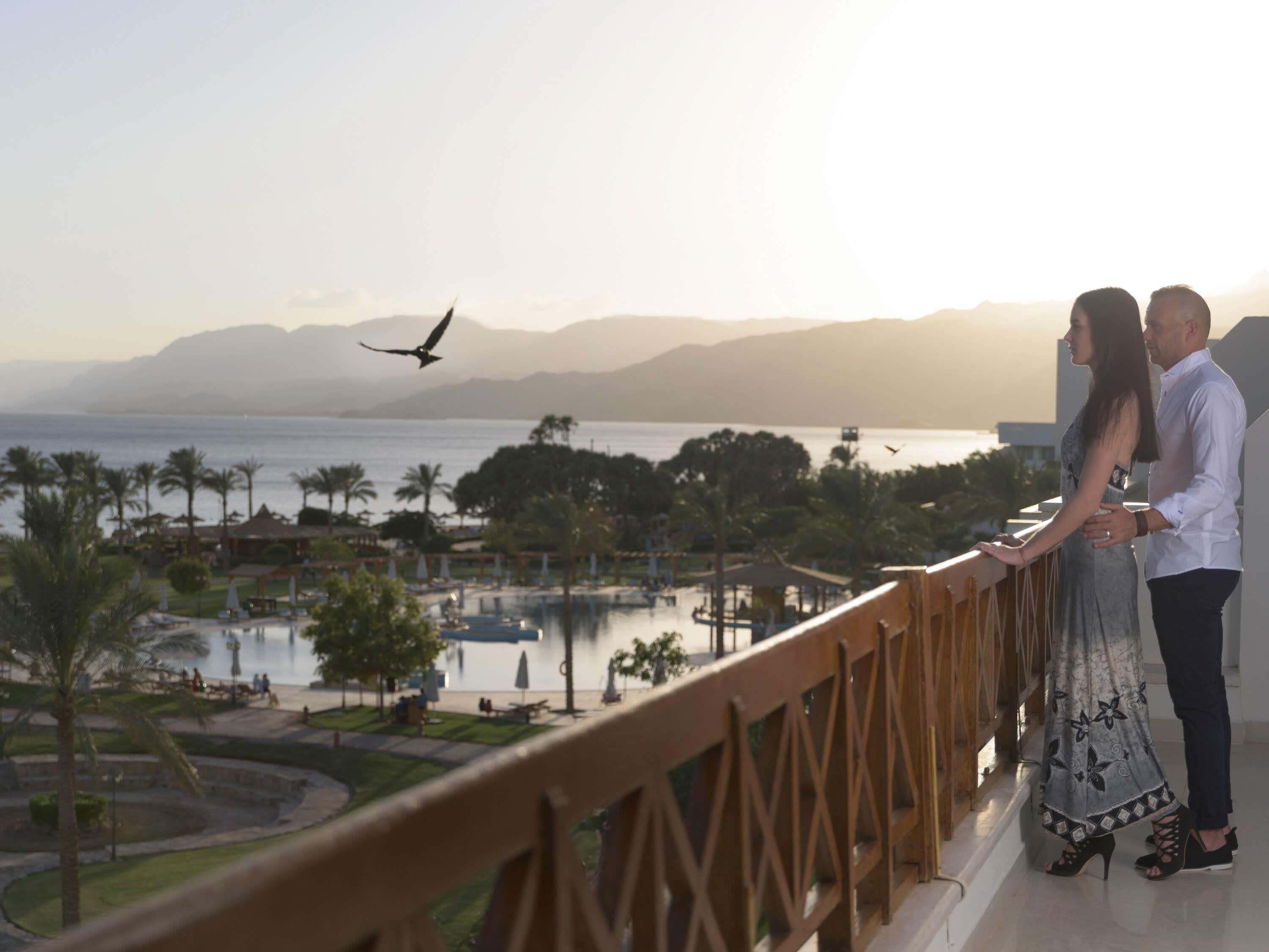 A couple standing on a balcony overlooking a pool and resort area with palm trees and mountains in the background.