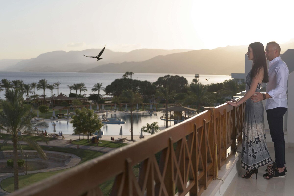 A couple standing on a balcony overlooking a pool and resort area with palm trees and mountains in the background.