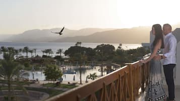 Um casal está de pé em uma varanda com vista para uma piscina e área de resort, com palmeiras e montanhas ao fundo.
