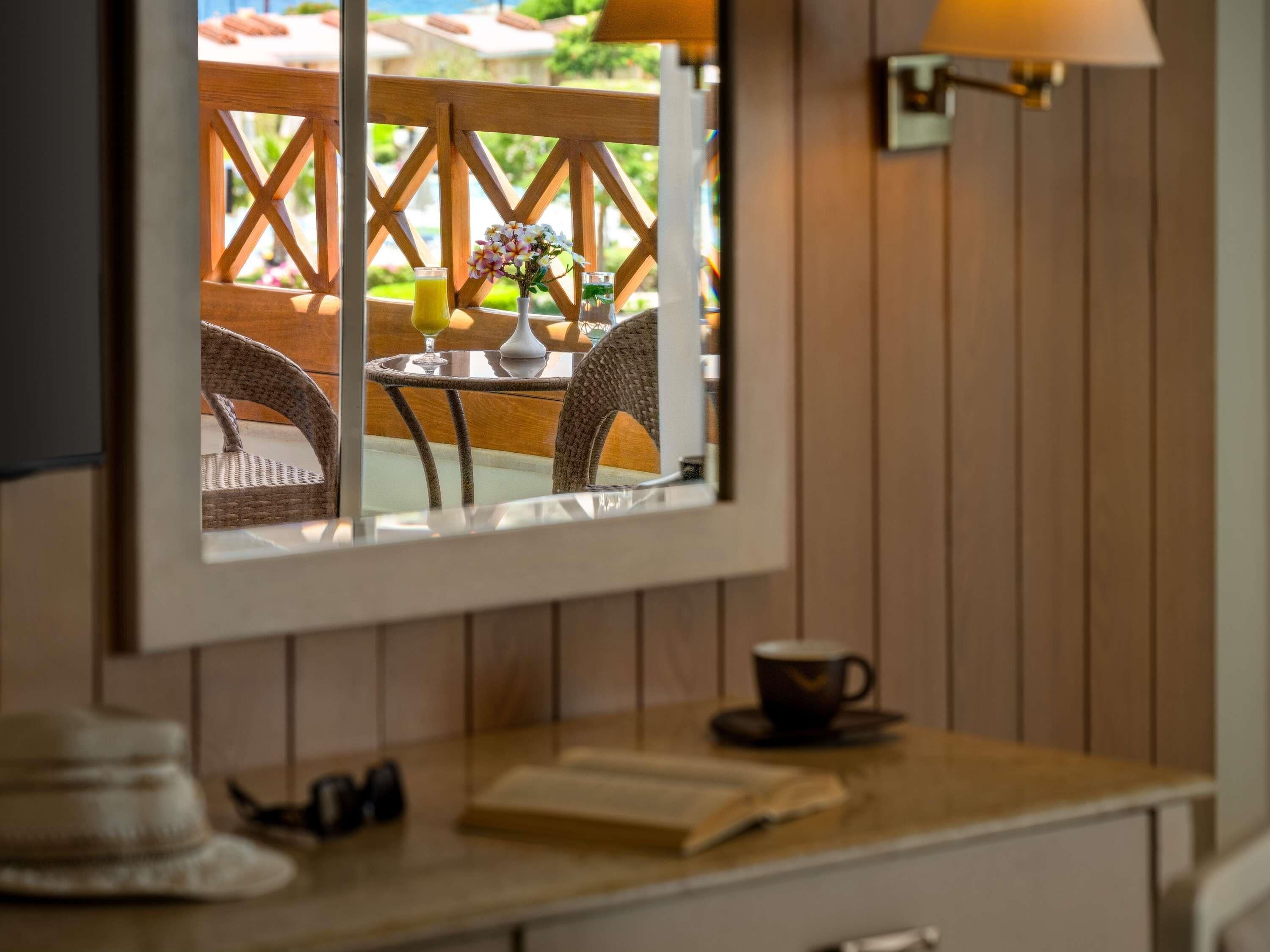 Ein Zimmer mit einer Holzwand, einem Spiegel, der einen Balkon mit Gartenmöbeln und einem Tisch zeigt, sowie einem kleinen Tisch mit Tasse und Buch.
