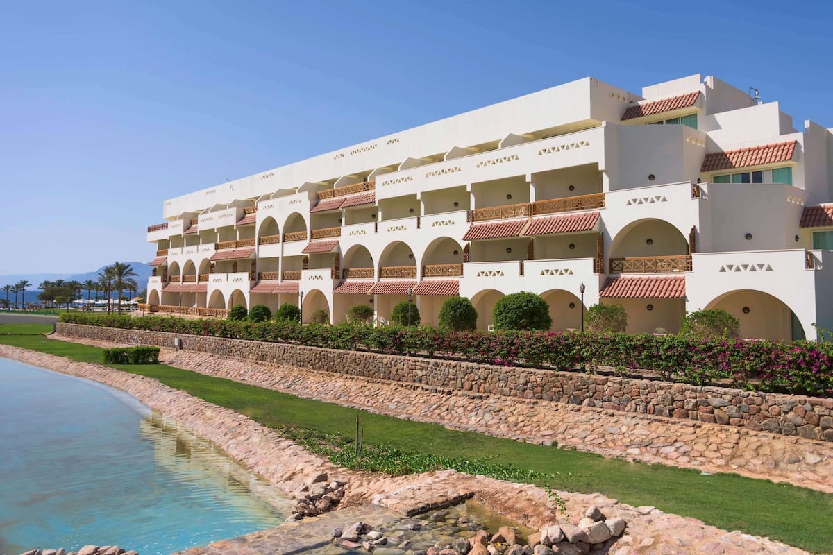 Un hotel de varios pisos con balcones, piscina y un muro de piedra.