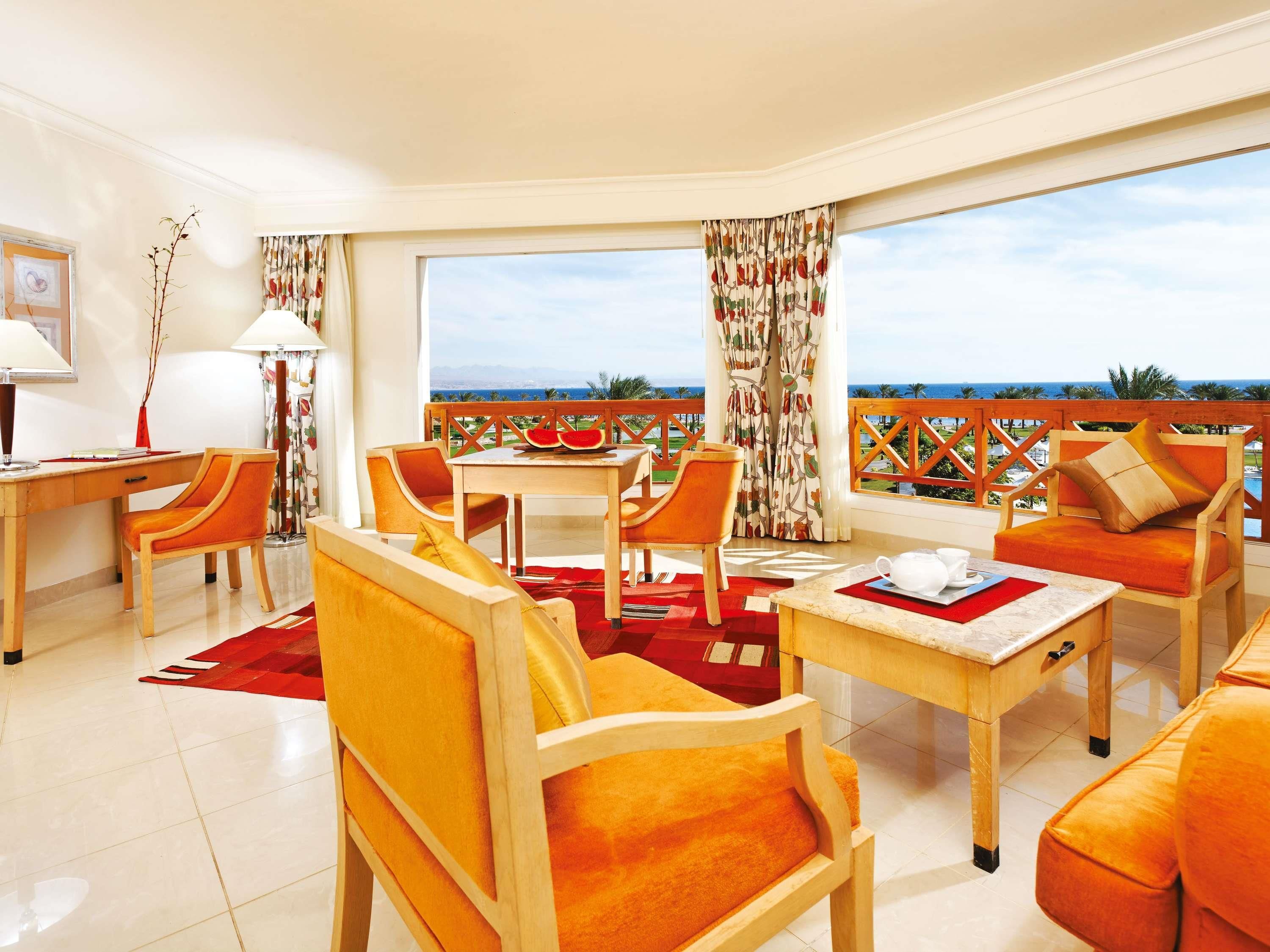 A modern hotel room with a balcony, a dining area, and a seating area with orange furniture.