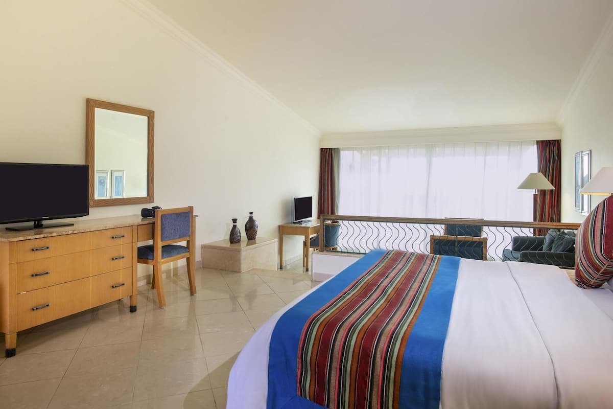 A hotel room with a bed, a desk with a TV, a chair, a small table, and a window with curtains.