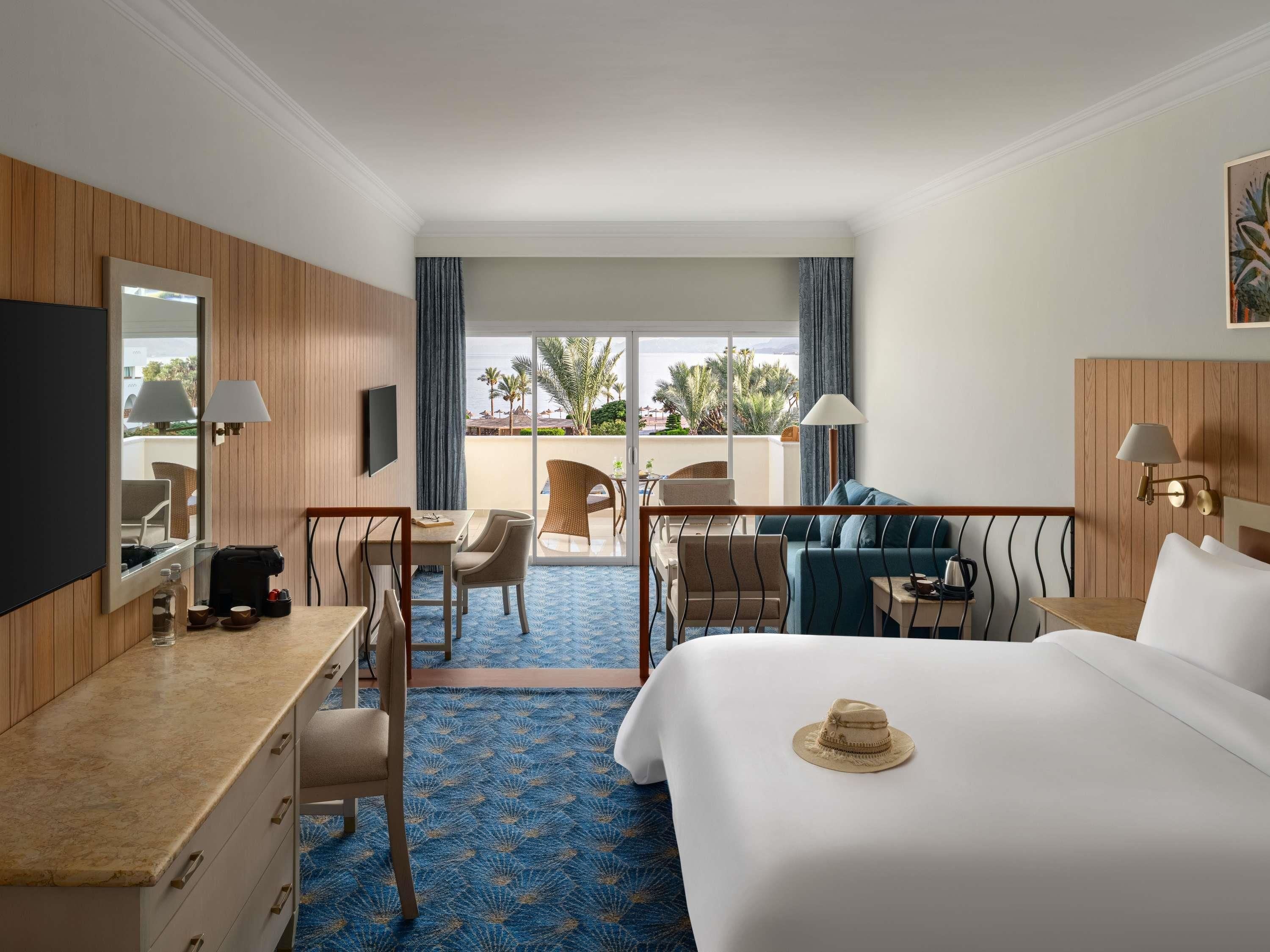 Habitación de hotel con cama, escritorio, sillas, televisor y balcón con vistas.