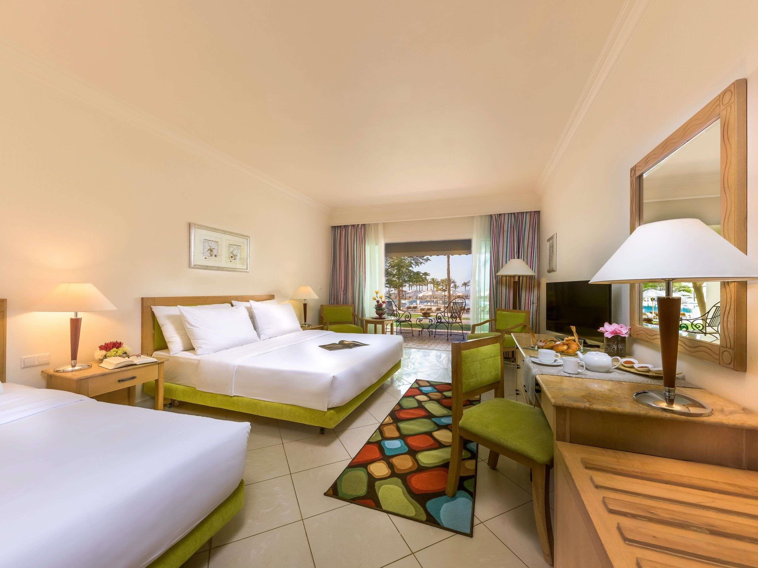 A hotel room with two beds, a desk, a chair, a TV, and a view of the outdoors.