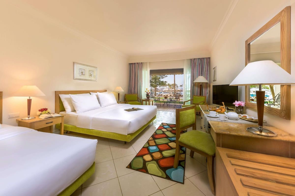 A hotel room with two beds, a desk, a chair, a TV, and a view of the outdoors.