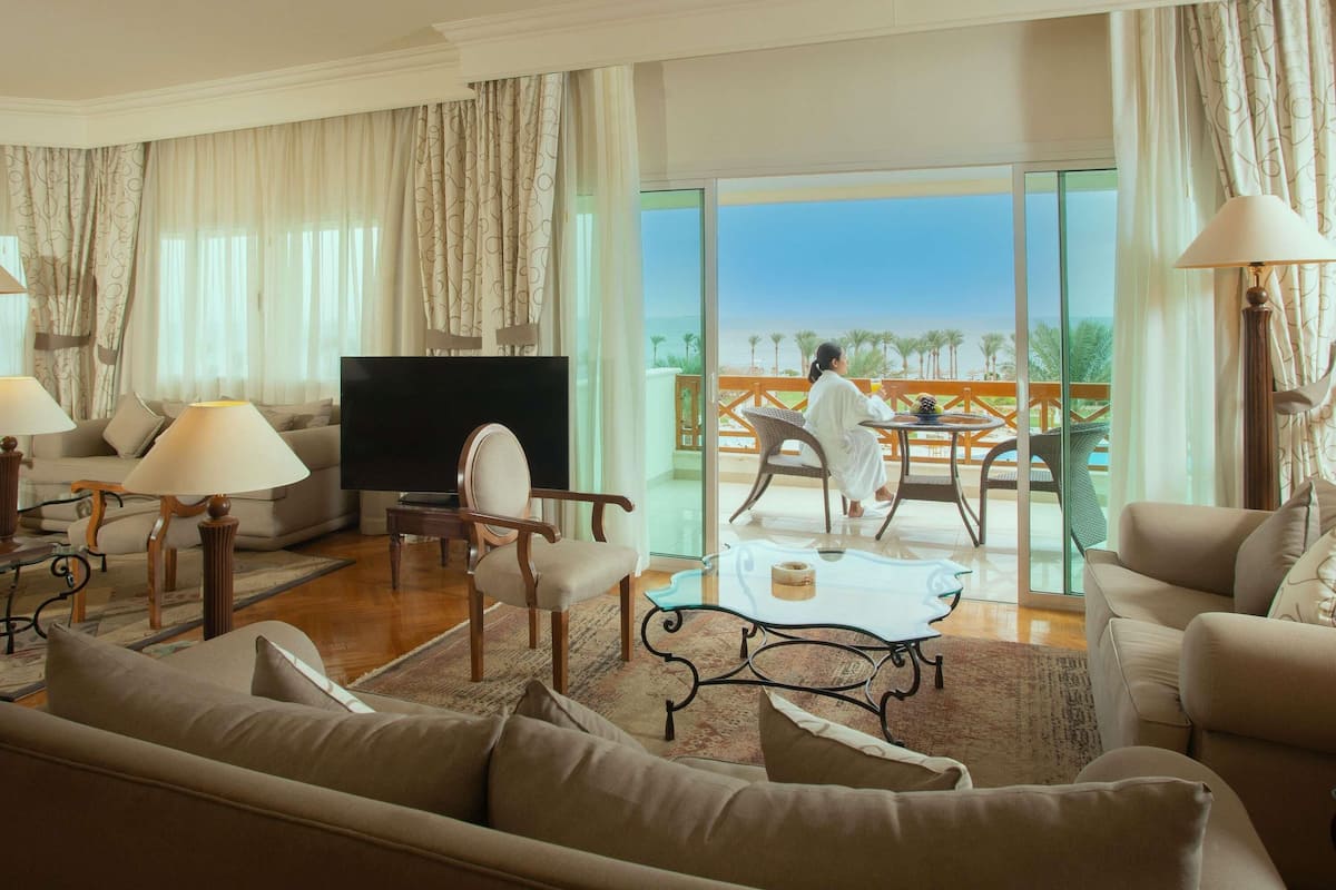 A spacious living room with a flat-screen TV, comfortable seating, and a balcony with a view of the beach.