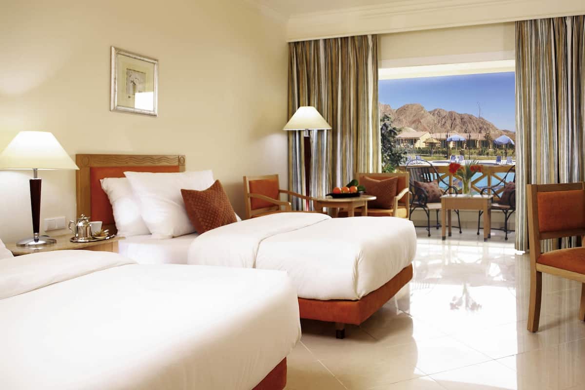 A hotel room with two beds, a dining area with a table and chairs, and a view of mountains through the window.