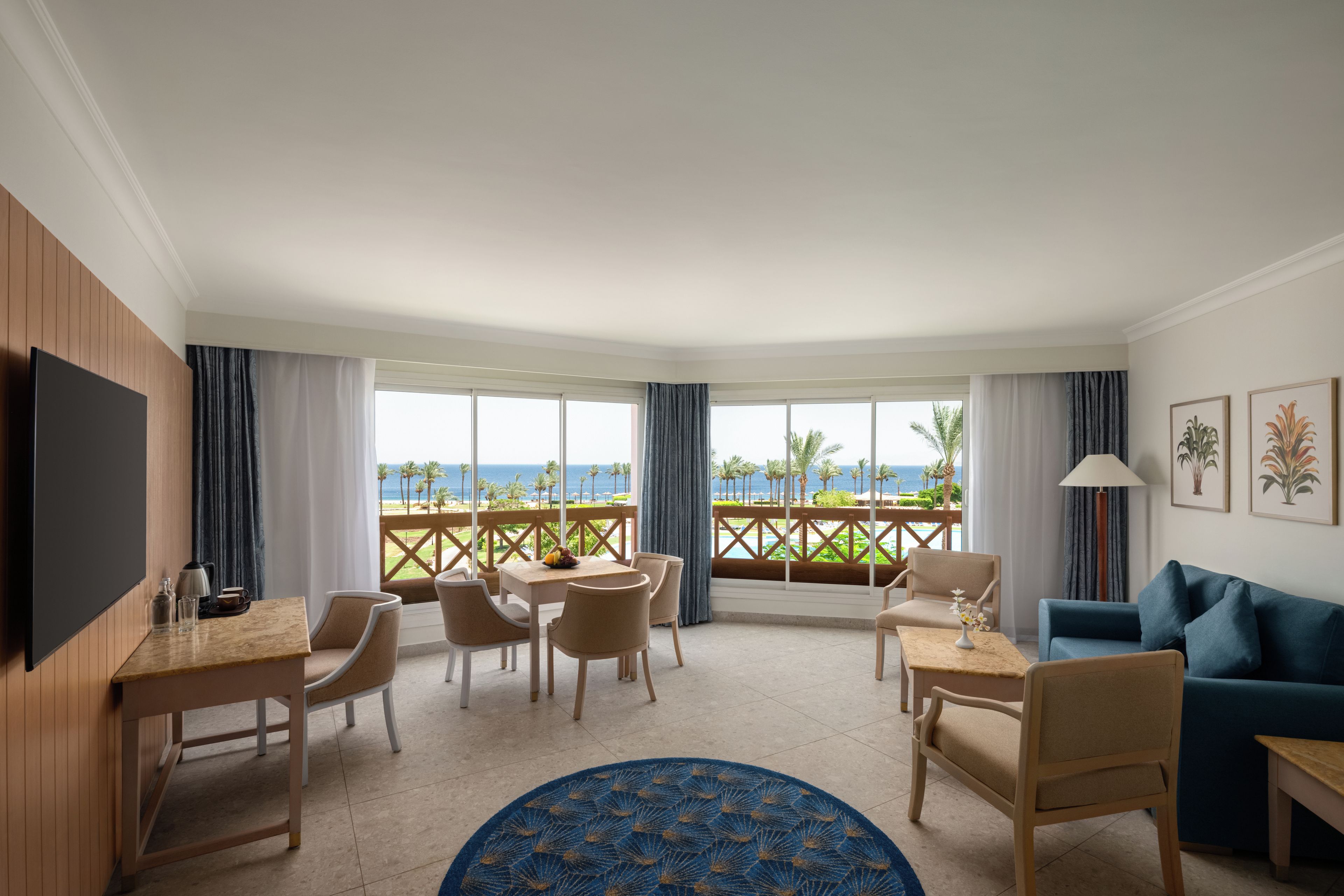 Habitación de hotel con televisión, zona de comedor con sillas y mesa, sofá, alfombra azul y balcón con vista al mar.