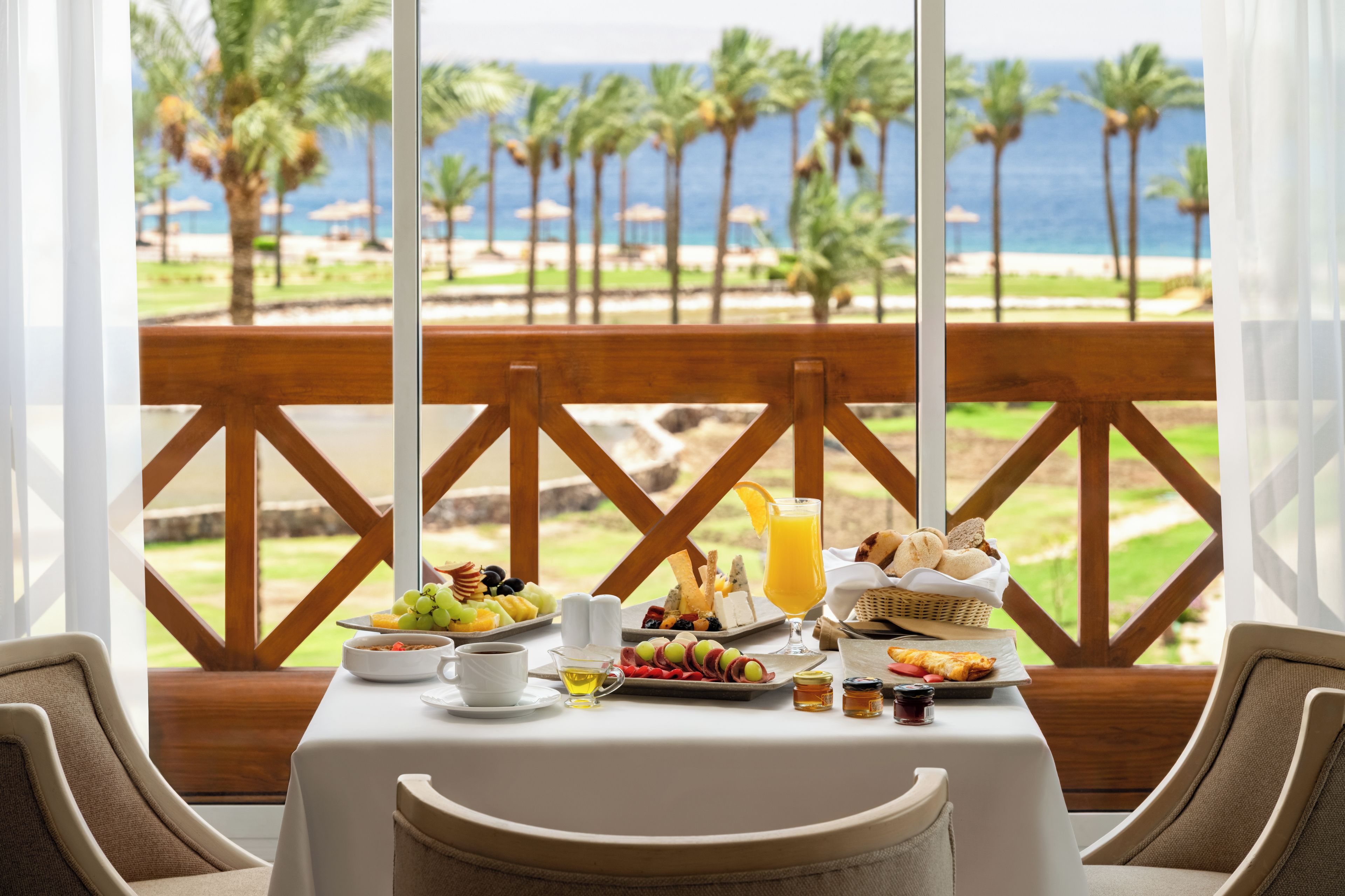 Une table à manger dressée avec des aliments pour le petit-déjeuner et un verre de jus d’orange, donnant sur une plage bordée de palmiers.
