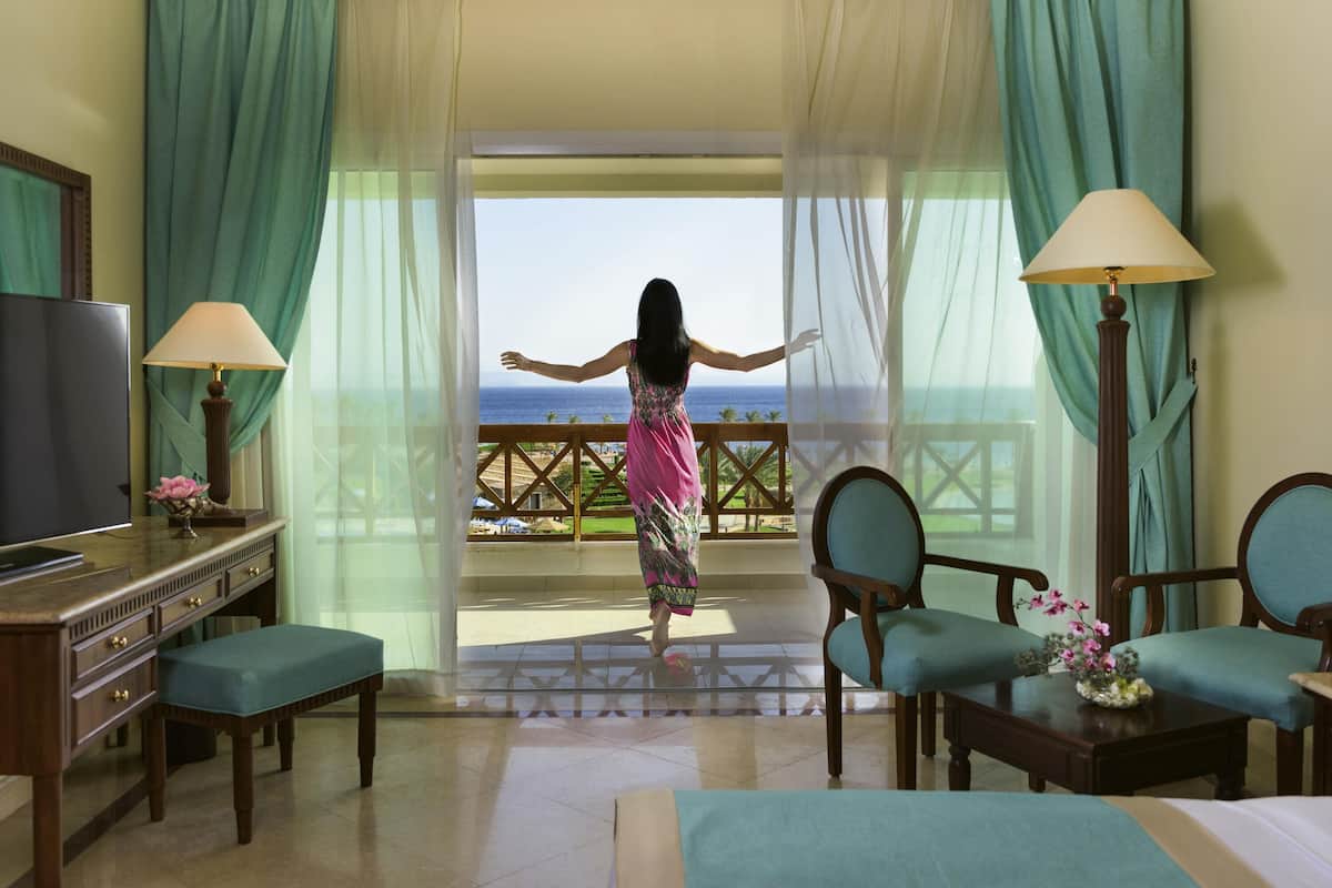 A room with a bed, a desk, two chairs, a TV, and a view of the ocean.