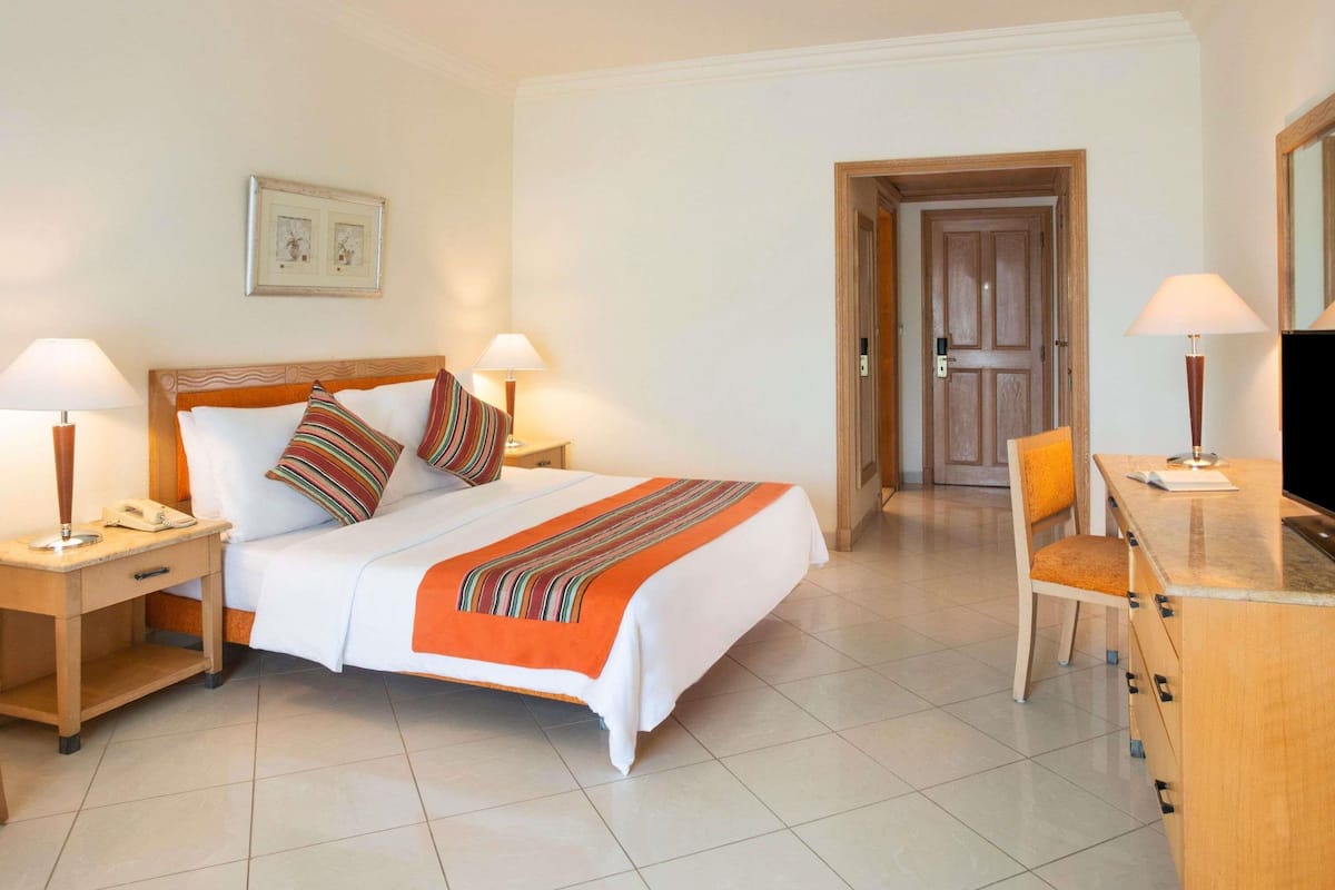 A hotel room with a bed, two bedside tables with lamps, a desk with a chair, and a door leading to another room.