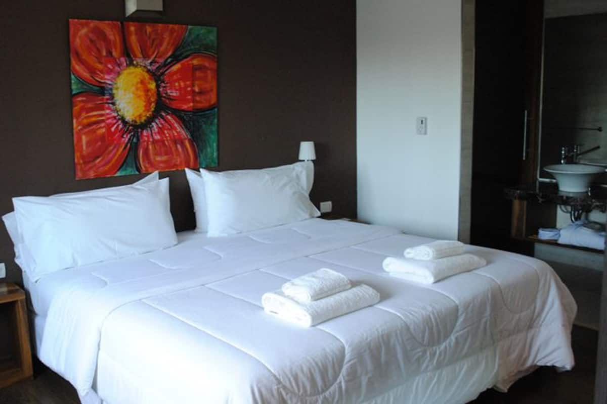Une chambre d’hôtel avec un grand lit, du linge de lit blanc, un tableau représentant une fleur accroché au mur et une salle de bain visible à travers une porte ouverte.