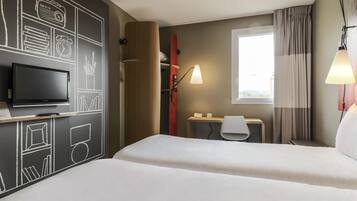 A hotel room with a bed, a desk with a chair, a TV, and a window with curtains.