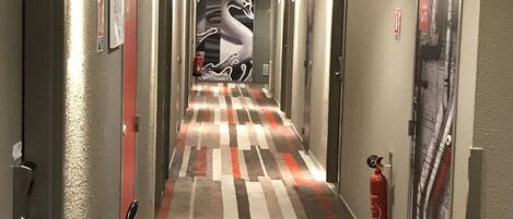 A hotel hallway with a patterned carpet, fire extinguisher, and a wall-mounted poster.