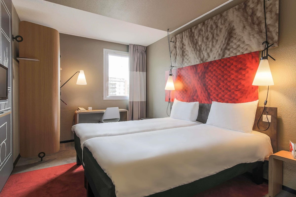 A hotel room with a large bed, a desk, a chair, a TV, and a window.