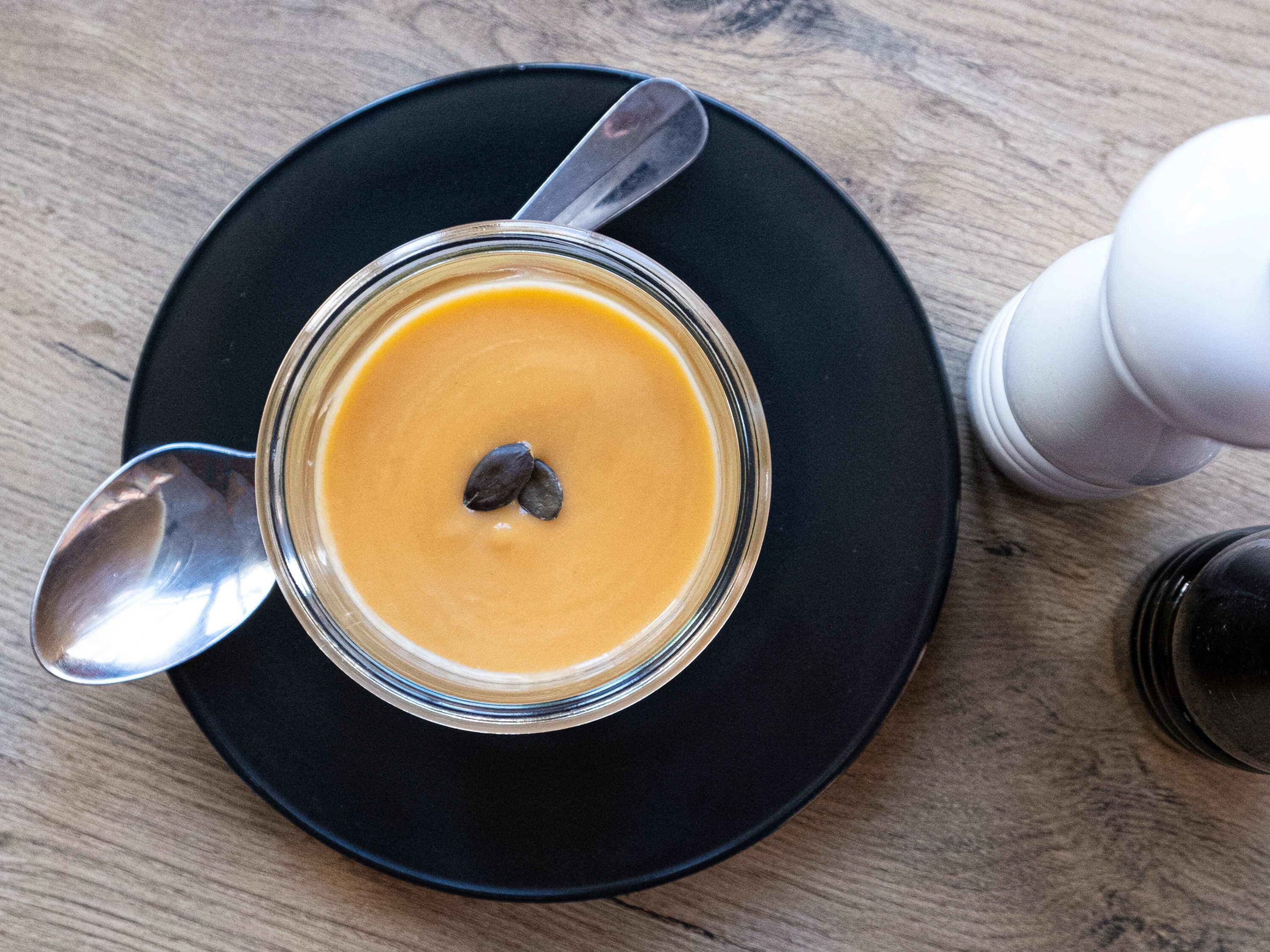 A glass bowl of soup on a black plate with a spoon, placed on a wooden table.