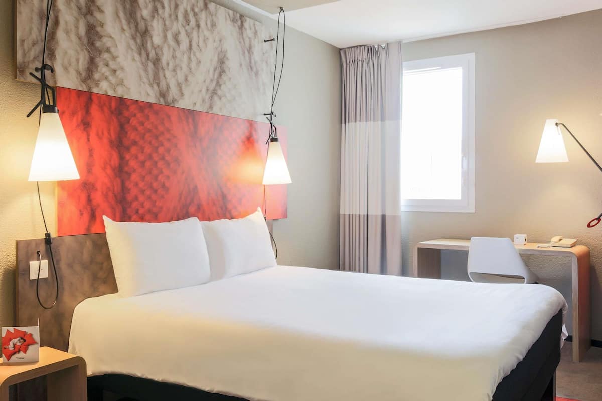 A hotel room with a large bed, two bedside lamps, a desk with a chair, a window with curtains, and a marble-patterned headboard.