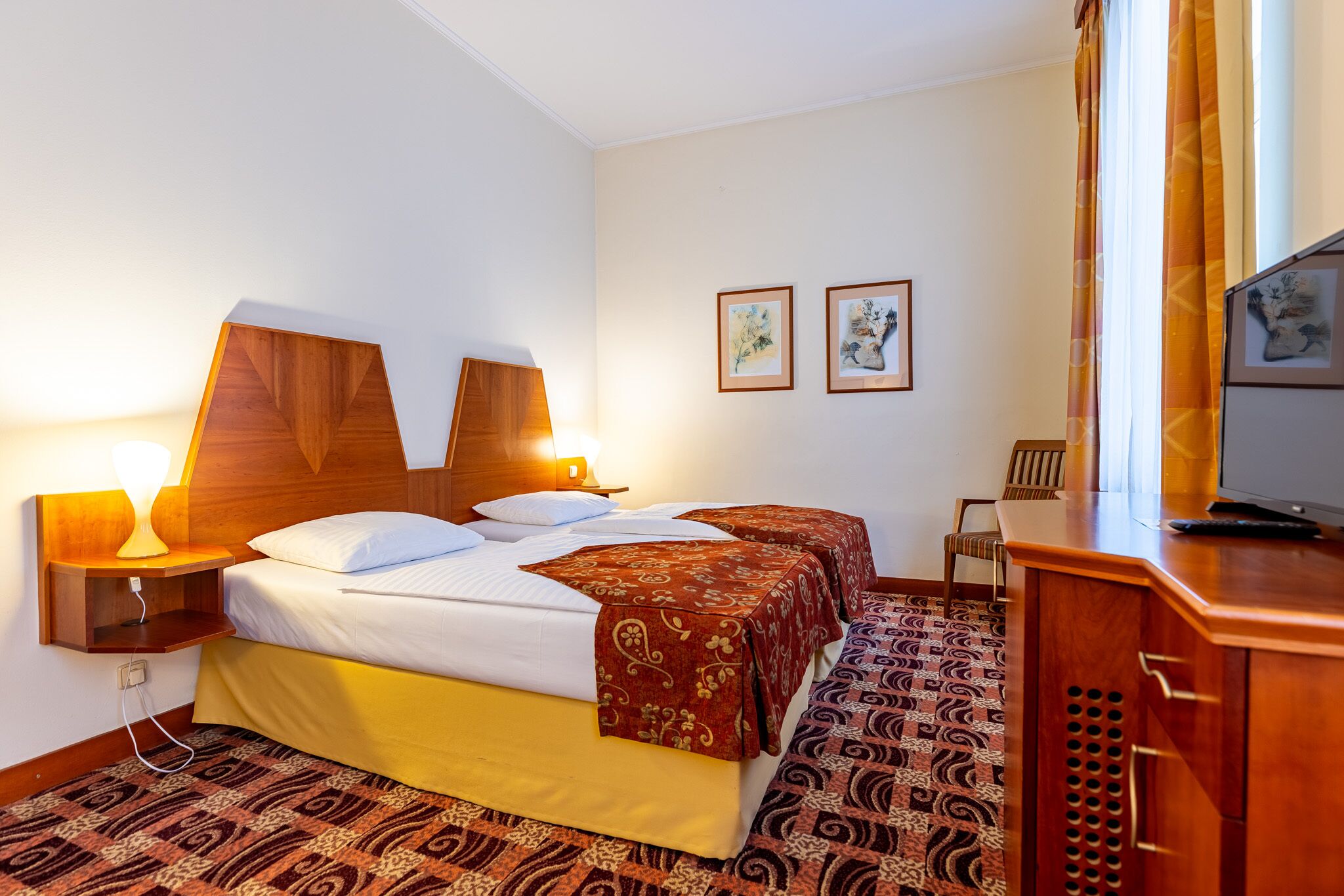 A hotel room with two beds, a wooden headboard, a desk with a television, and patterned carpet.