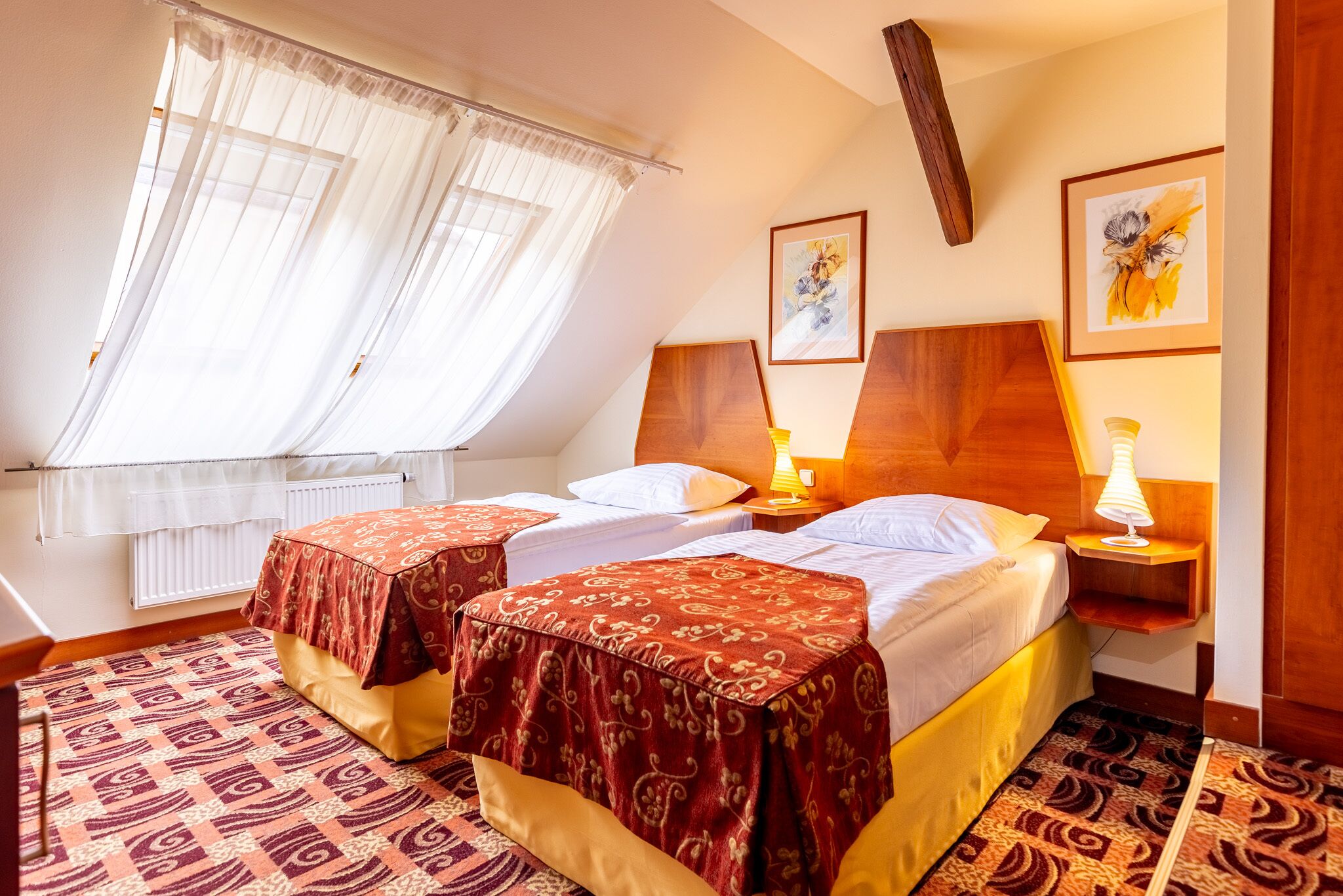 Quarto de hotel com duas camas, uma janela com cortinas translúcidas, cabeceira de madeira e carpete estampado.