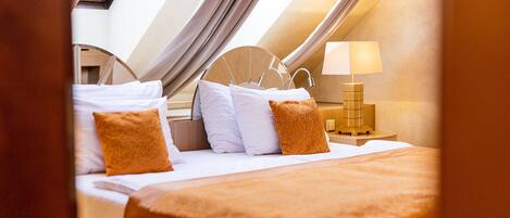 Un dormitorio con tragaluces, una cama con ropa blanca y almohadas anaranjadas, una mesita de noche con lámpara y un cabecero de madera.