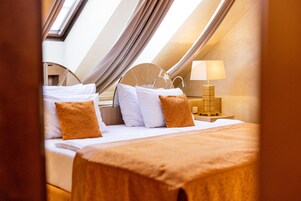 Un dormitorio con tragaluces, una cama con ropa blanca y almohadas anaranjadas, una mesita de noche con lámpara y un cabecero de madera.
