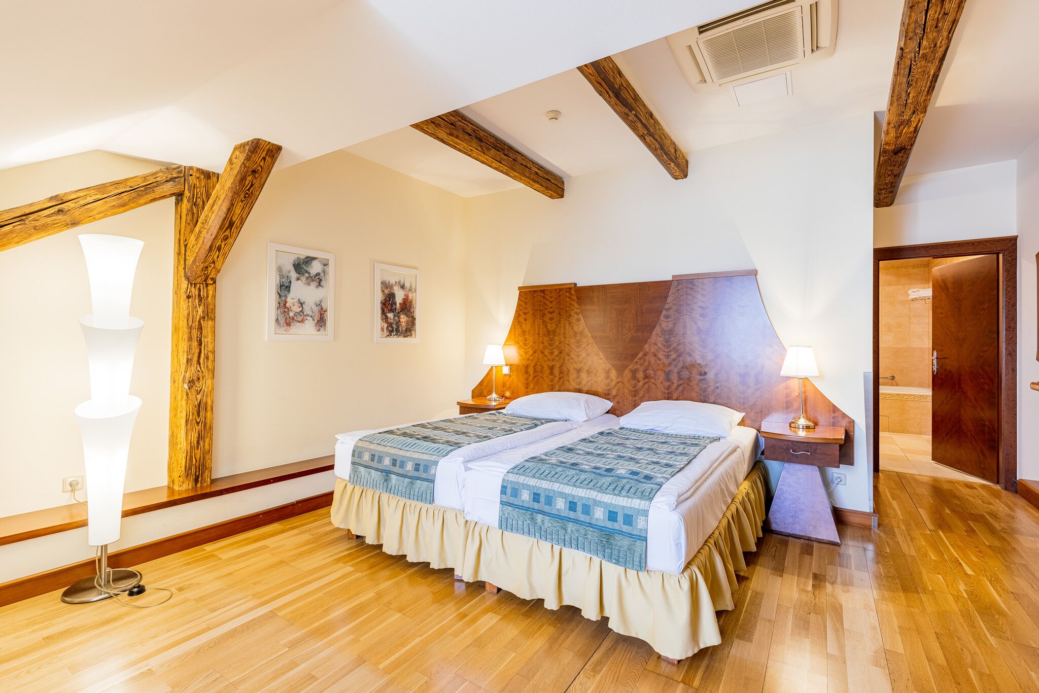 Quarto com teto de madeira, cama com colcha estampada, duas luminárias de cabeceira e mesa de cabeceira de madeira.